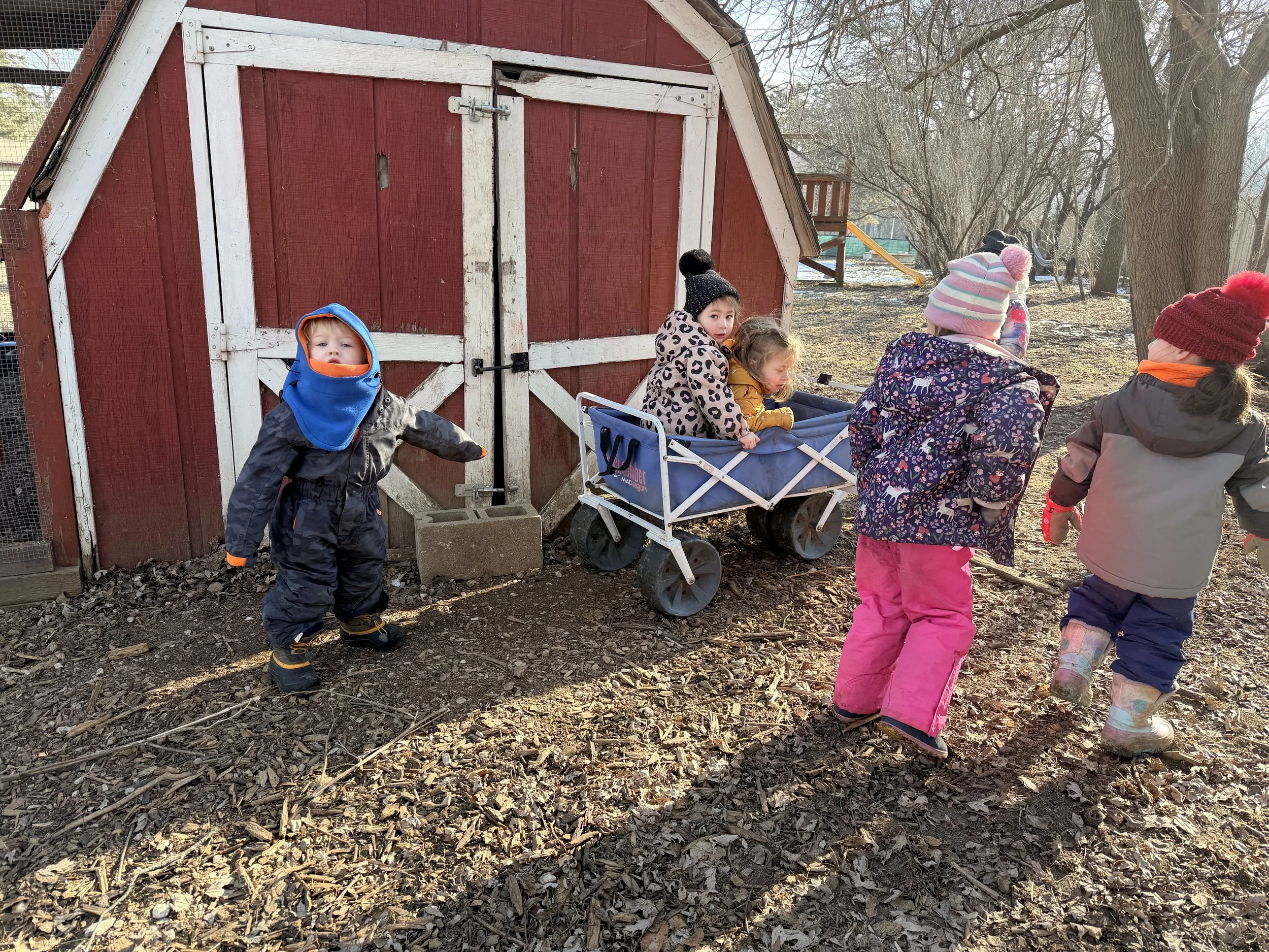 Children dressed warmly playing outdoors near a red wooden barn, with some children sitting and others standing, on a sunny day with leaf-covered ground and leafless trees.