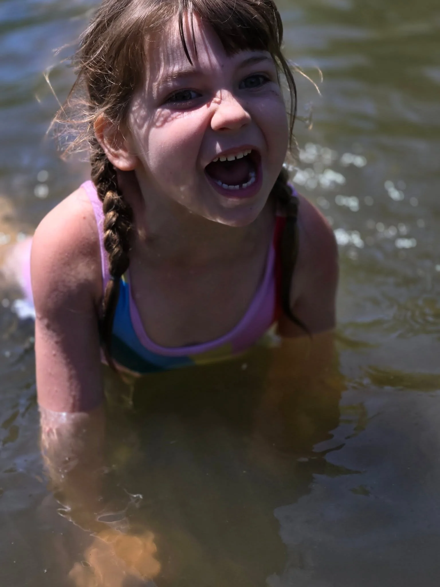 A young girl with braided hair is in shallow water, smiling with her mouth open.