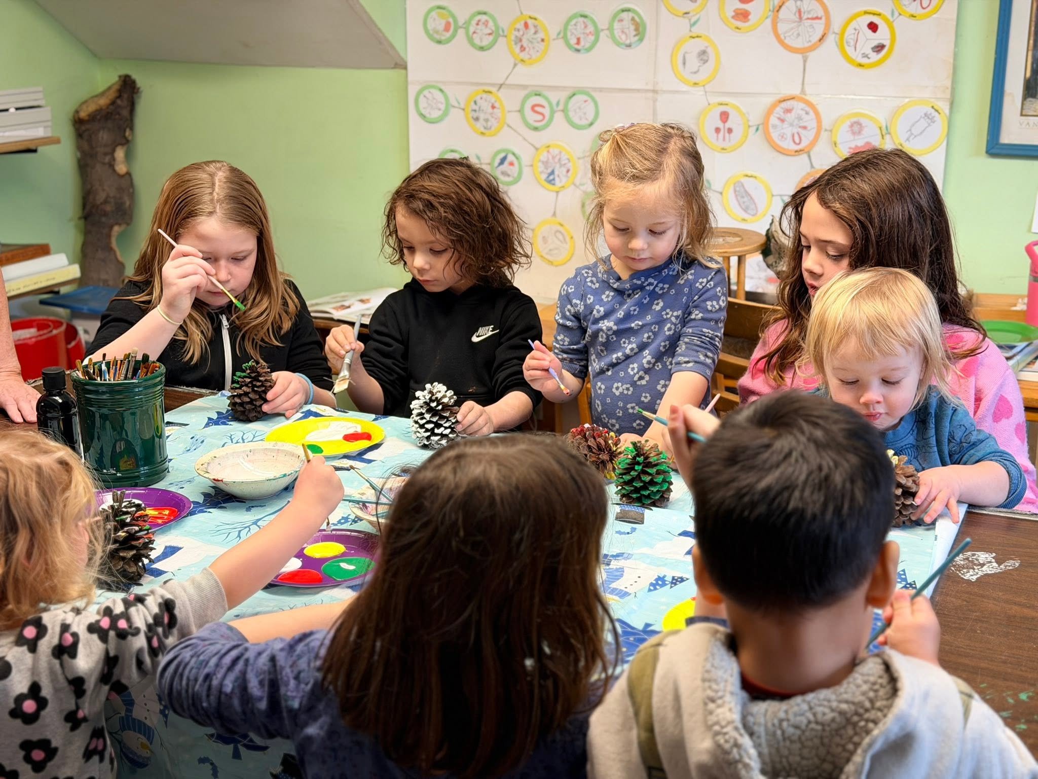Children participating in a craft activity, painting pinecones at a table in a colorful classroom with educational posters on the wall.