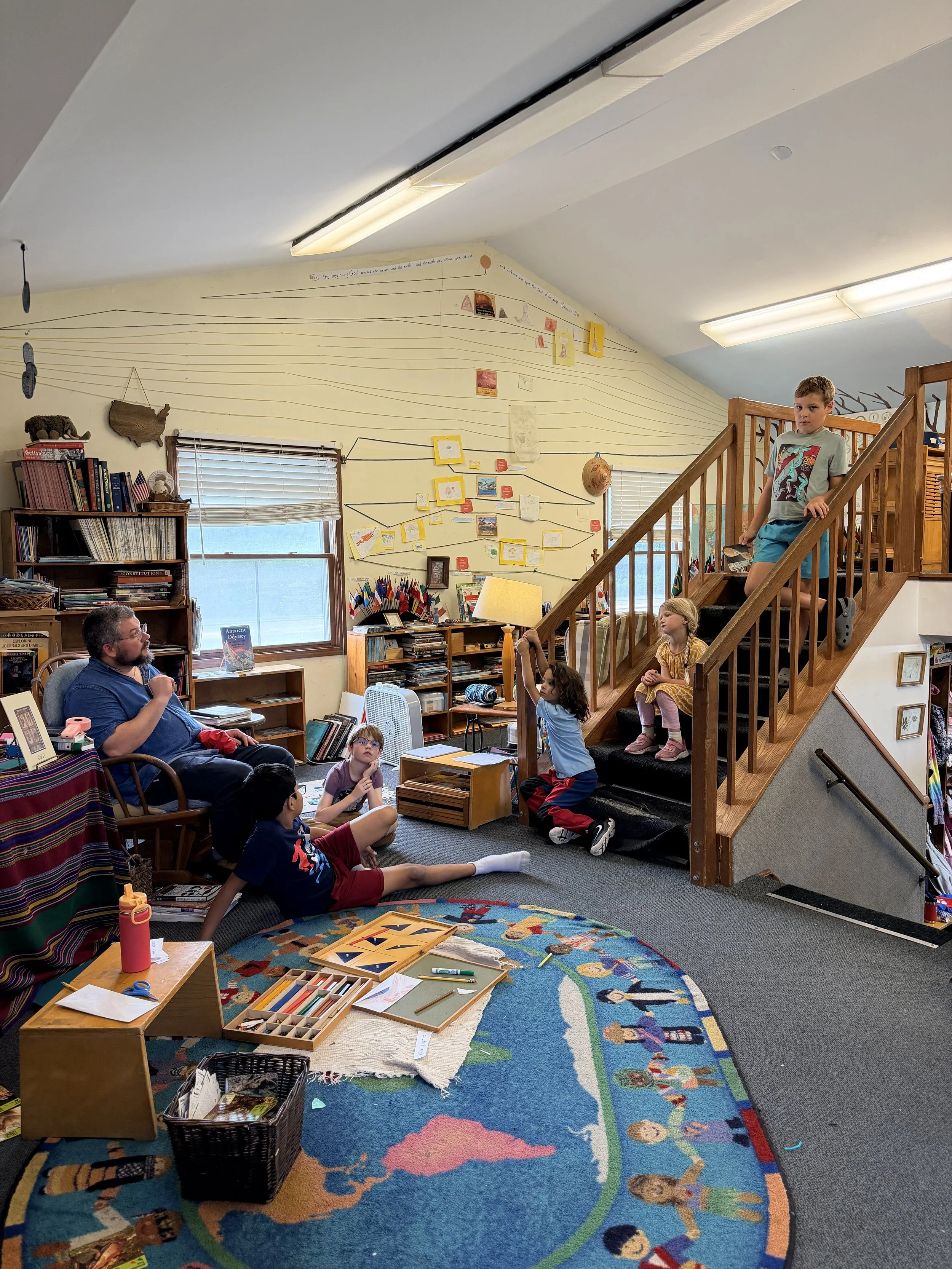 Interior of a classroom or community room with children and adults gathered on the floor and stairs, engaged in activities. A colorful rug with children's illustrations covers the floor, and shelves filled with books and educational materials are vis