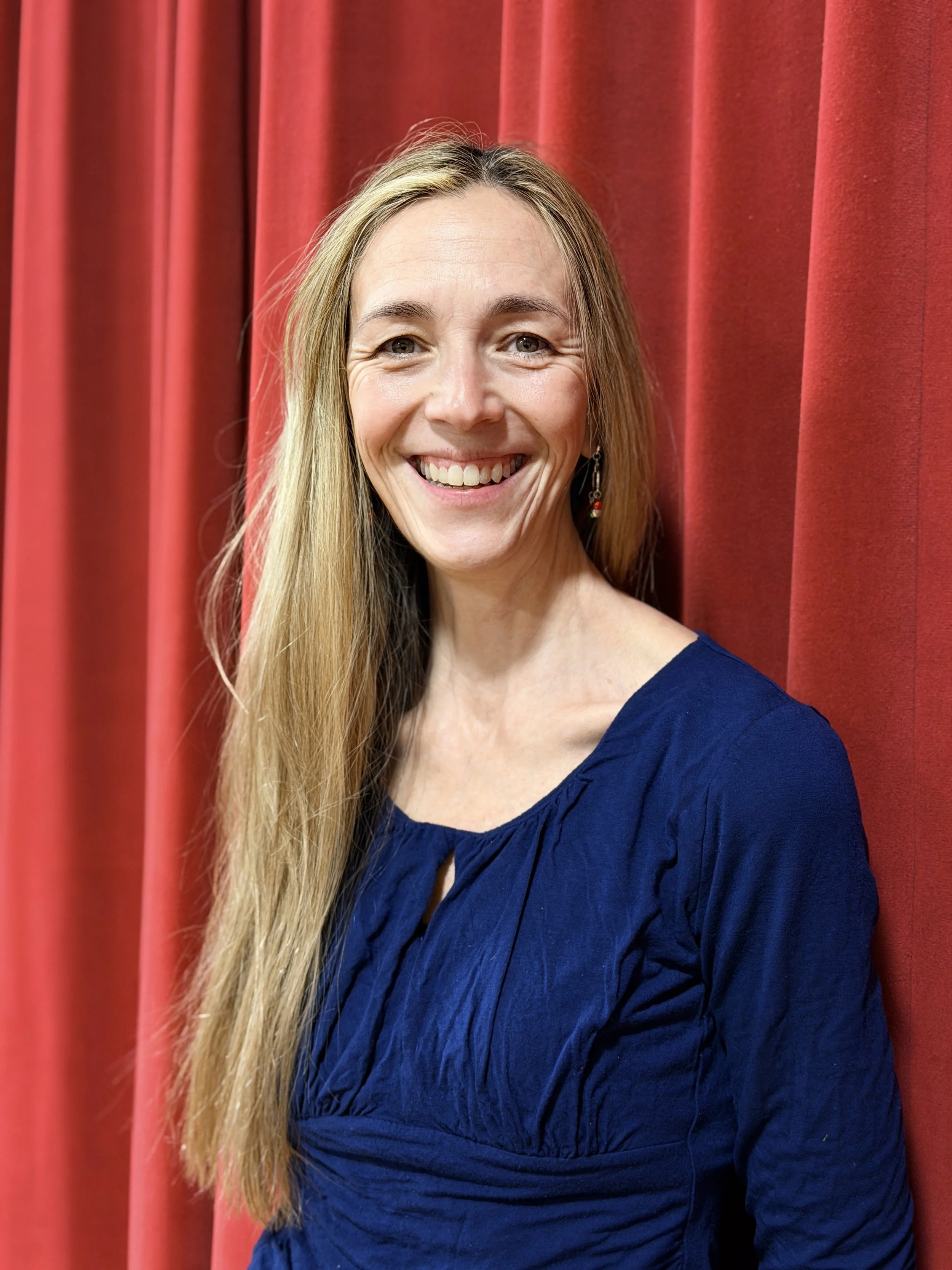 A woman with long blonde hair, wearing a blue dress, smiling, standing in front of red curtains.