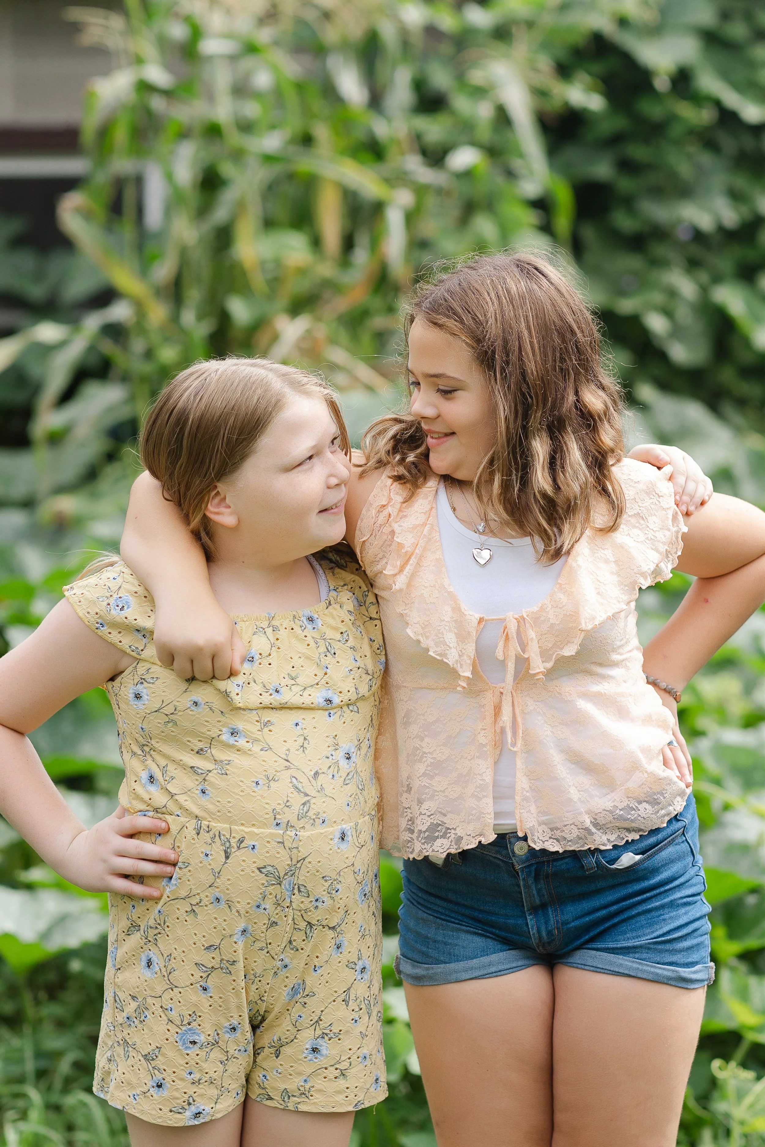 Two girls standing outdoors, smiling, with their arms around each other, in a green garden or farm setting.
