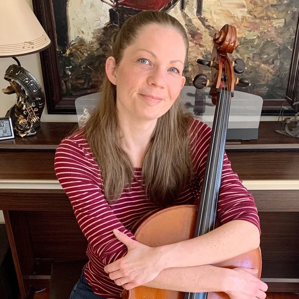 A woman with long brown hair and fair skin, wearing a red and white striped shirt, holds a cello in front of a piano and artwork in the background.