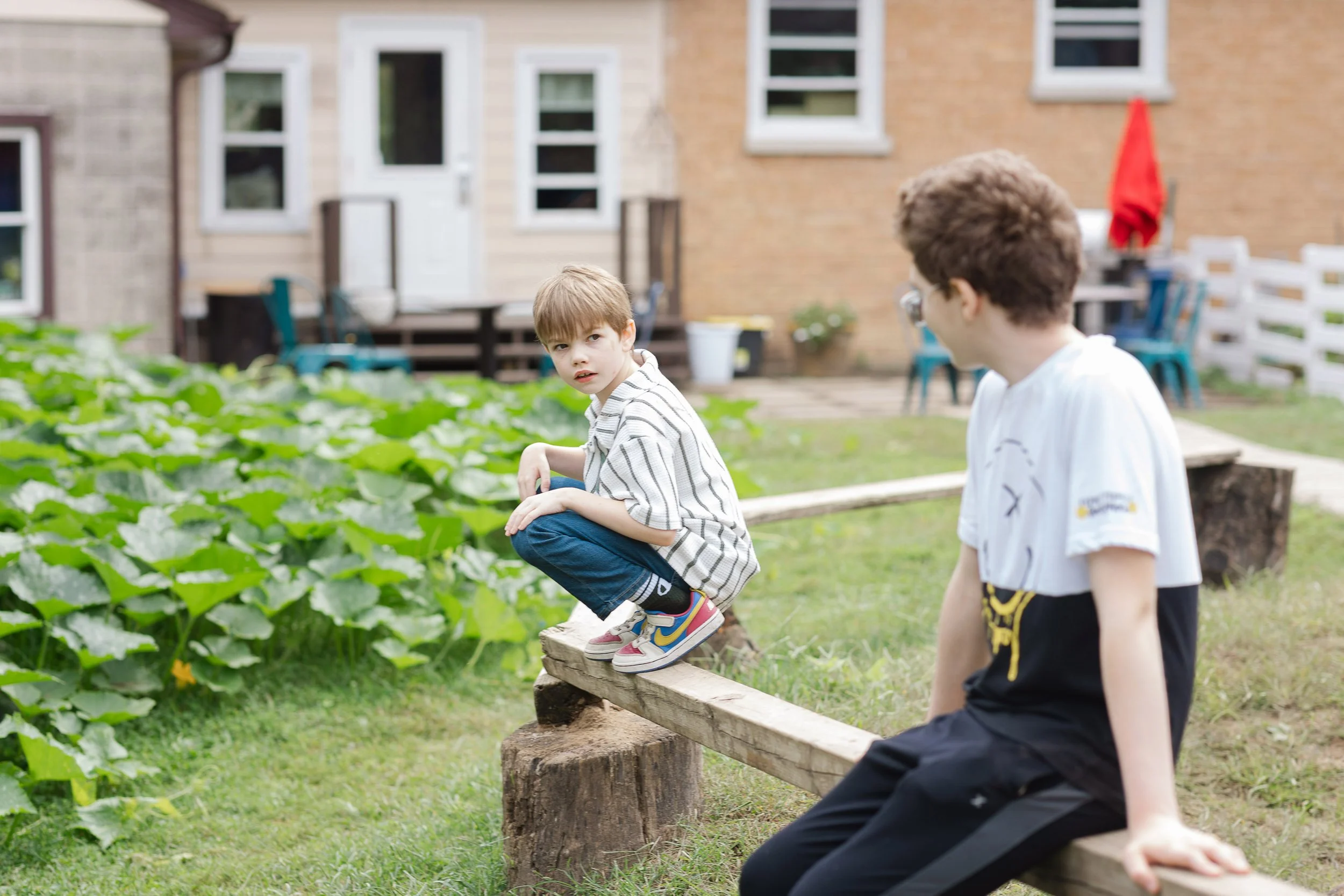 Two young boys sit on a wooden bench in a backyard garden, with one boy looking towards the camera and the other boy facing away. The boy facing the camera has light brown hair and is wearing a striped shirt and colorful sneakers. The other boy has c