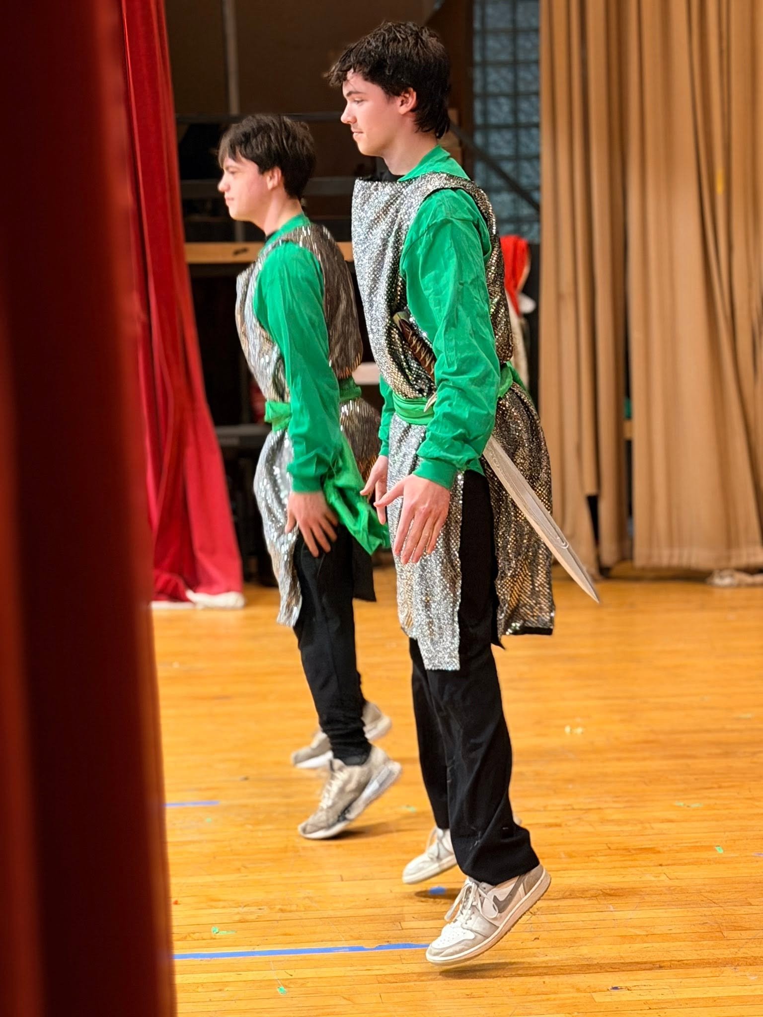 Two young men are dancing in a gymnasium with a polished wooden floor. They are dressed in shiny silver and green costumes with sneakers, and are positioned closely next to each other, performing synchronized dance moves.