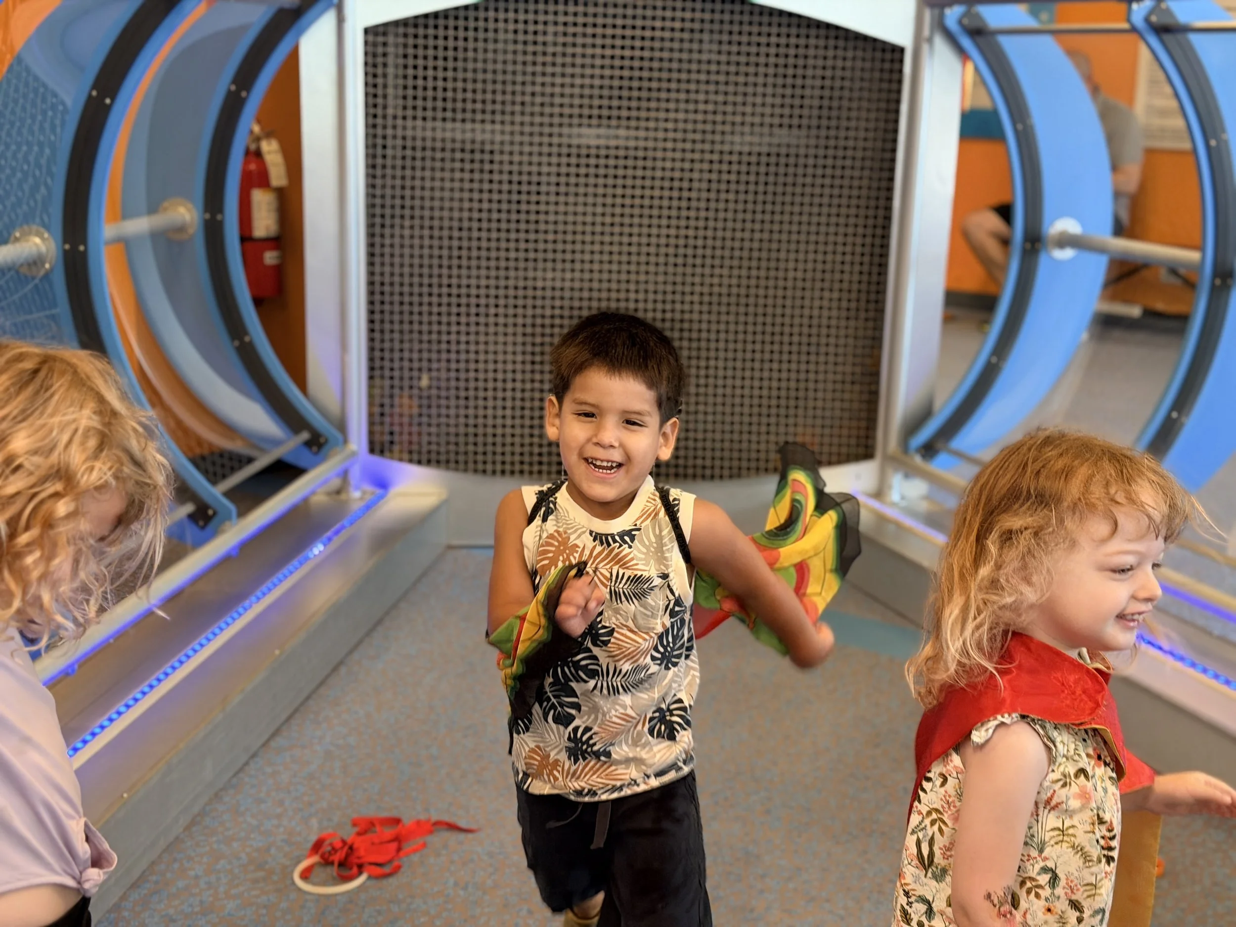 Smiling young boy wearing a tropical print shirt holding a rainbow-colored backpack while standing in an arcade or amusement center. Other children are partially visible around him.