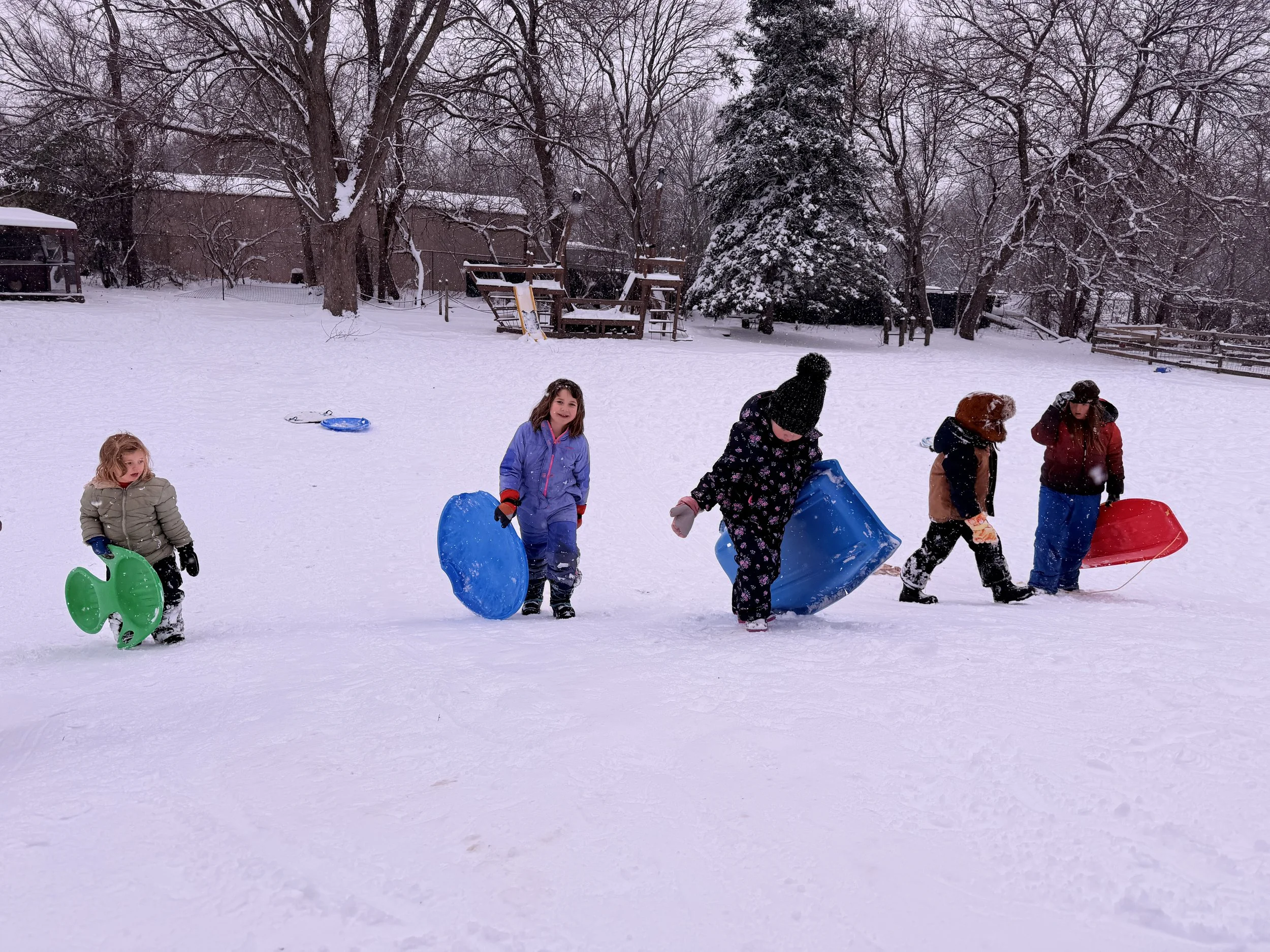 Children playing in a snowy backyard, holding sleds and wearing winter clothing, with snow-covered trees and a wooden structure in the background.