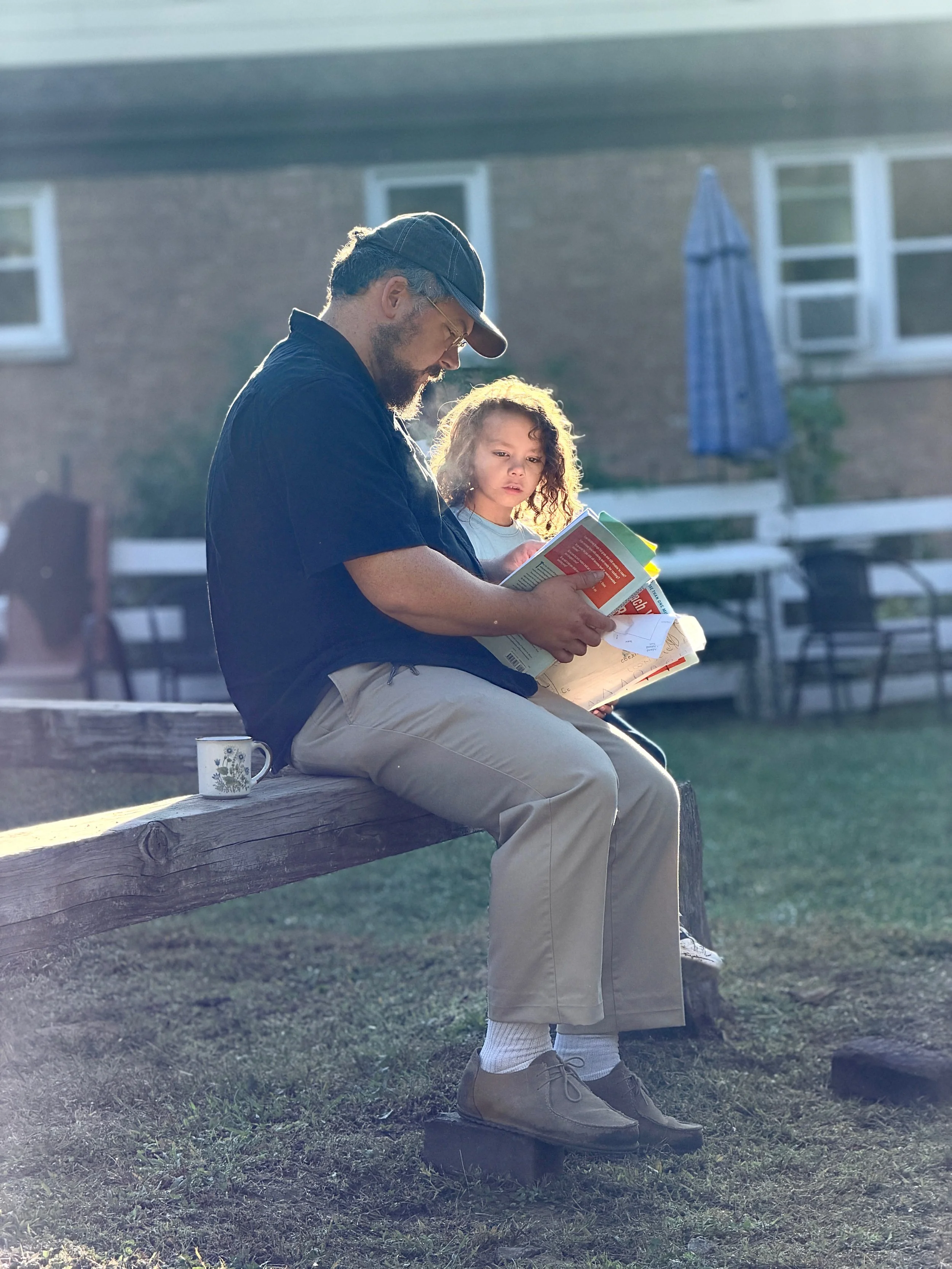 A man and a young girl are sitting on a wooden bench outdoors, reading a book together. The man is wearing a black shirt, beige pants, and a dark baseball cap, while the girl has curly hair and is dressed in a light blue top and beige pants. There is