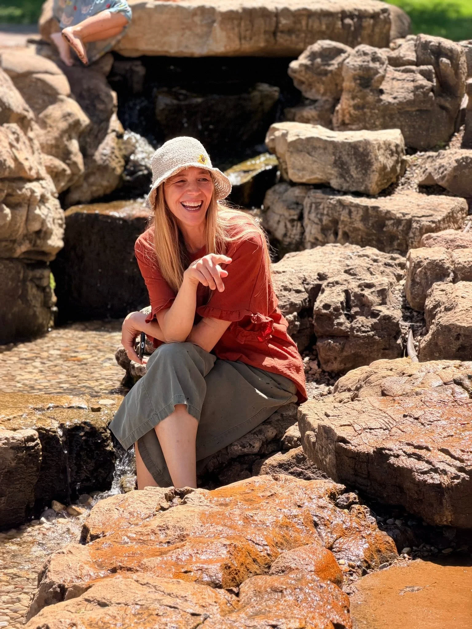 A woman with long blonde hair, wearing a beige sun hat, orange top, and gray pants, sitting on rocks near a small stream with rocks and water, smiling and pointing at the camera, in a sunny outdoor setting.