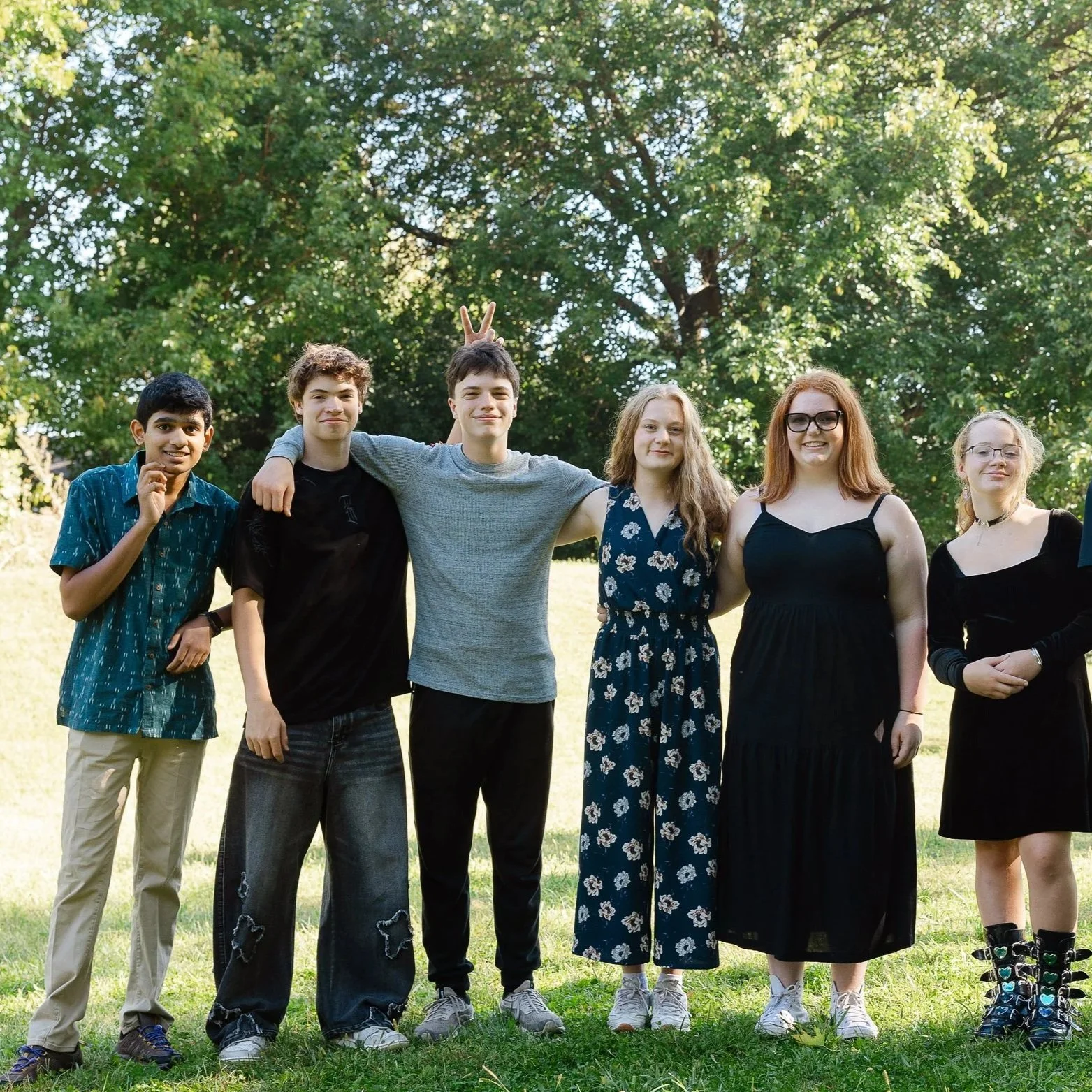 Group of six teenagers standing outdoors on grass with trees in the background, smiling and posing for a photo.