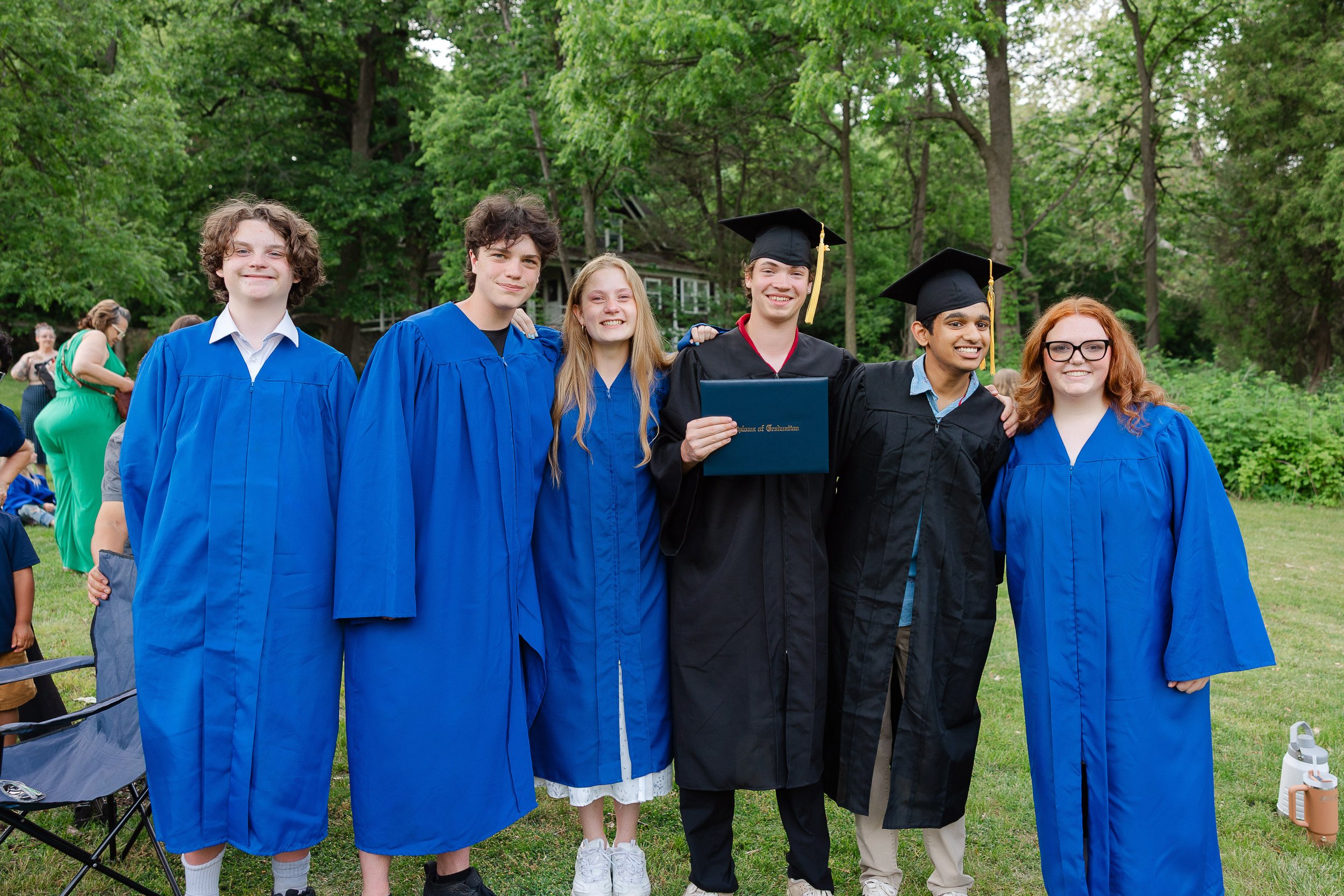 Group of six young graduates wearing caps and gowns, standing outdoors in a park, smiling for a photo.