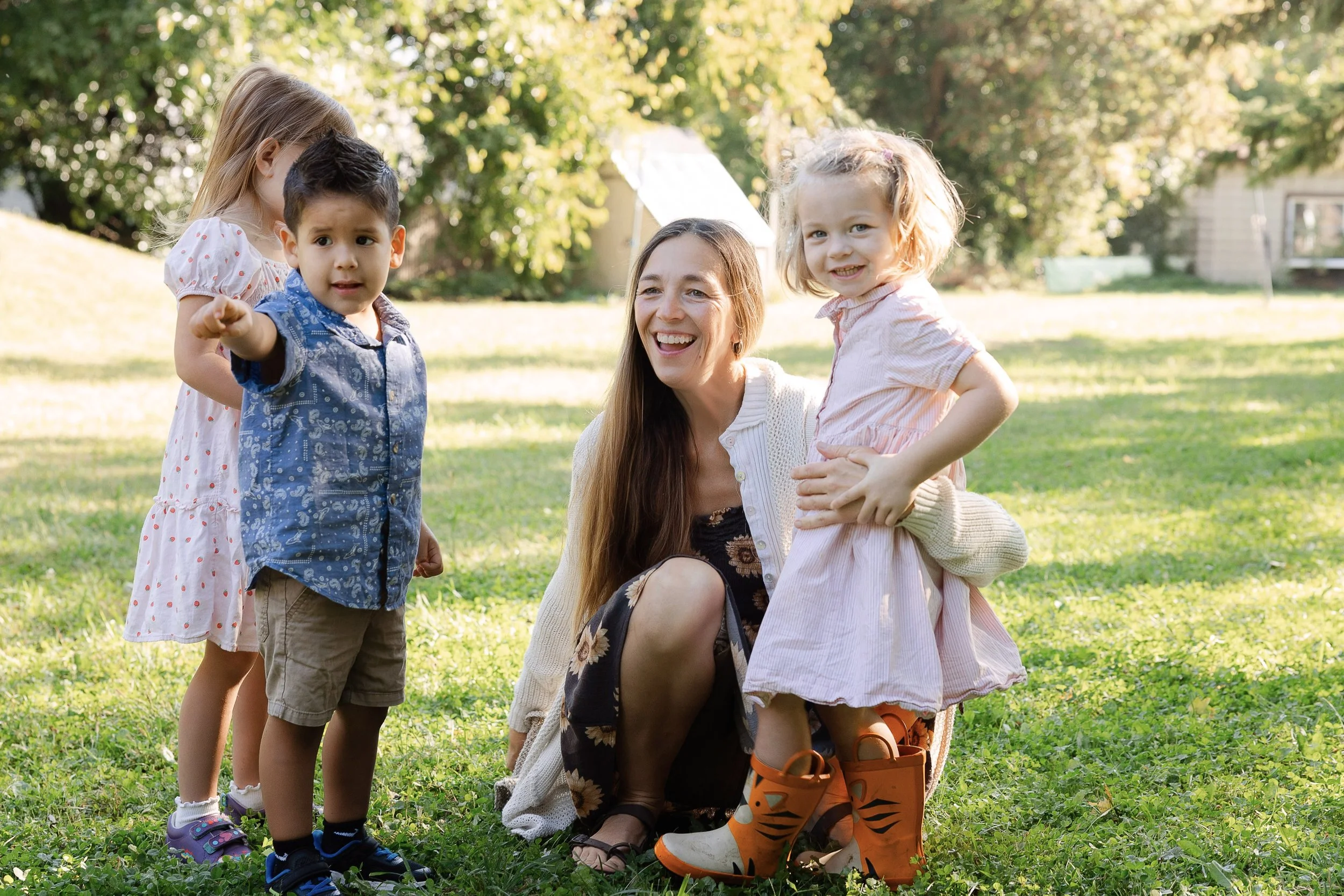 A woman crouches outdoors with four children, one pointing at the camera. The scene is sunny with green trees and grass in the background.