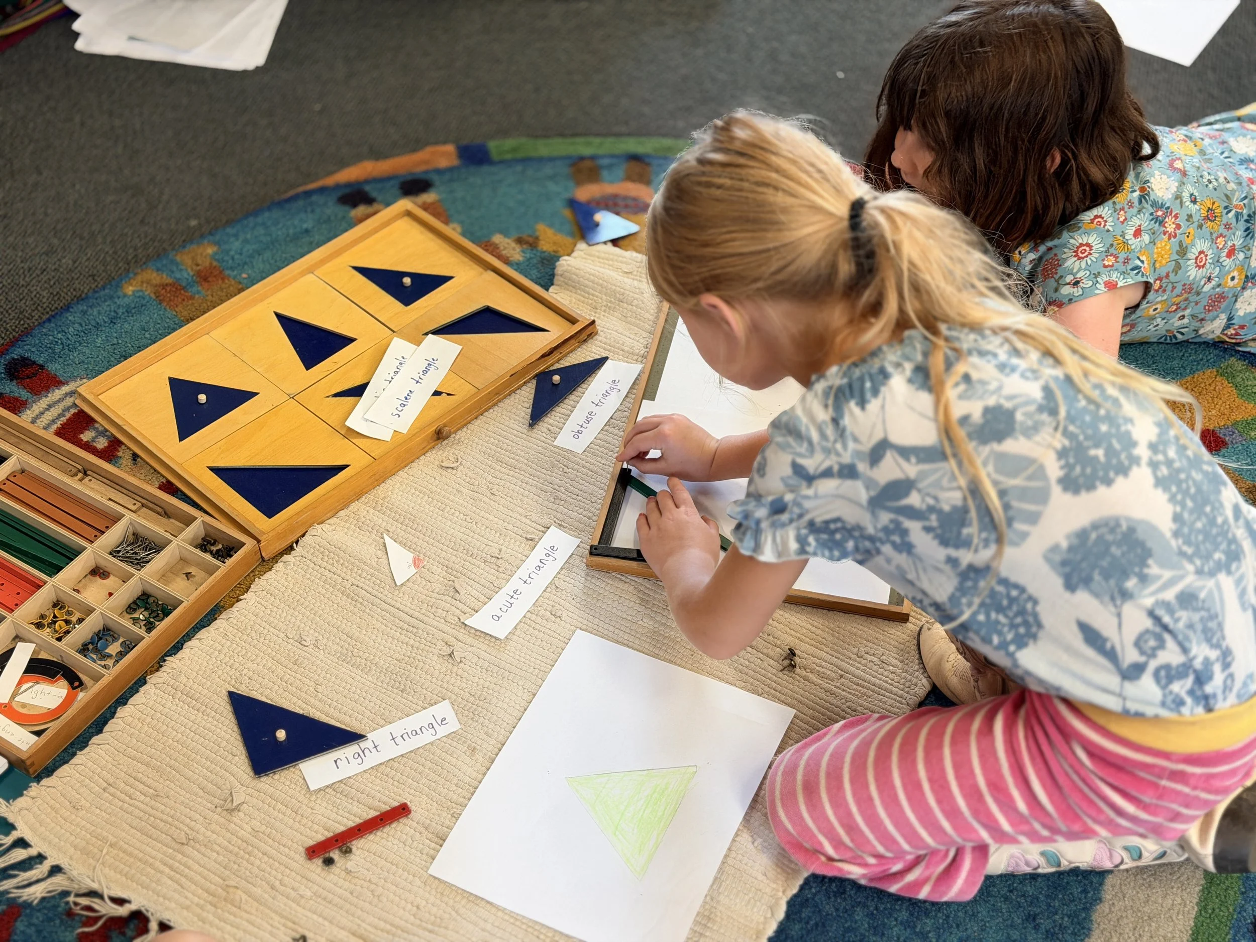 Two young girls sitting on a rug playing with geometric puzzle pieces and labeled cards, including words like 'acute triangle' and 'right triangle'.