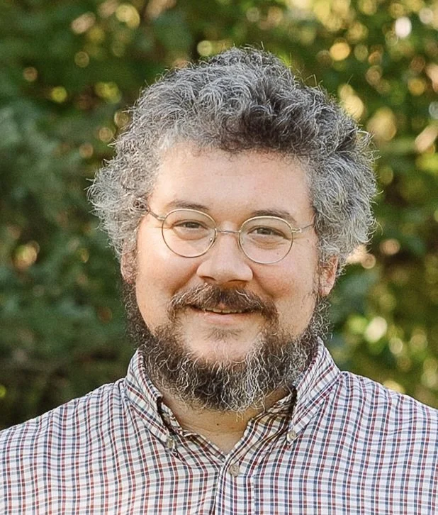 A man with gray curly hair and glasses, smiling outdoors with trees in the background, wearing a checkered shirt.