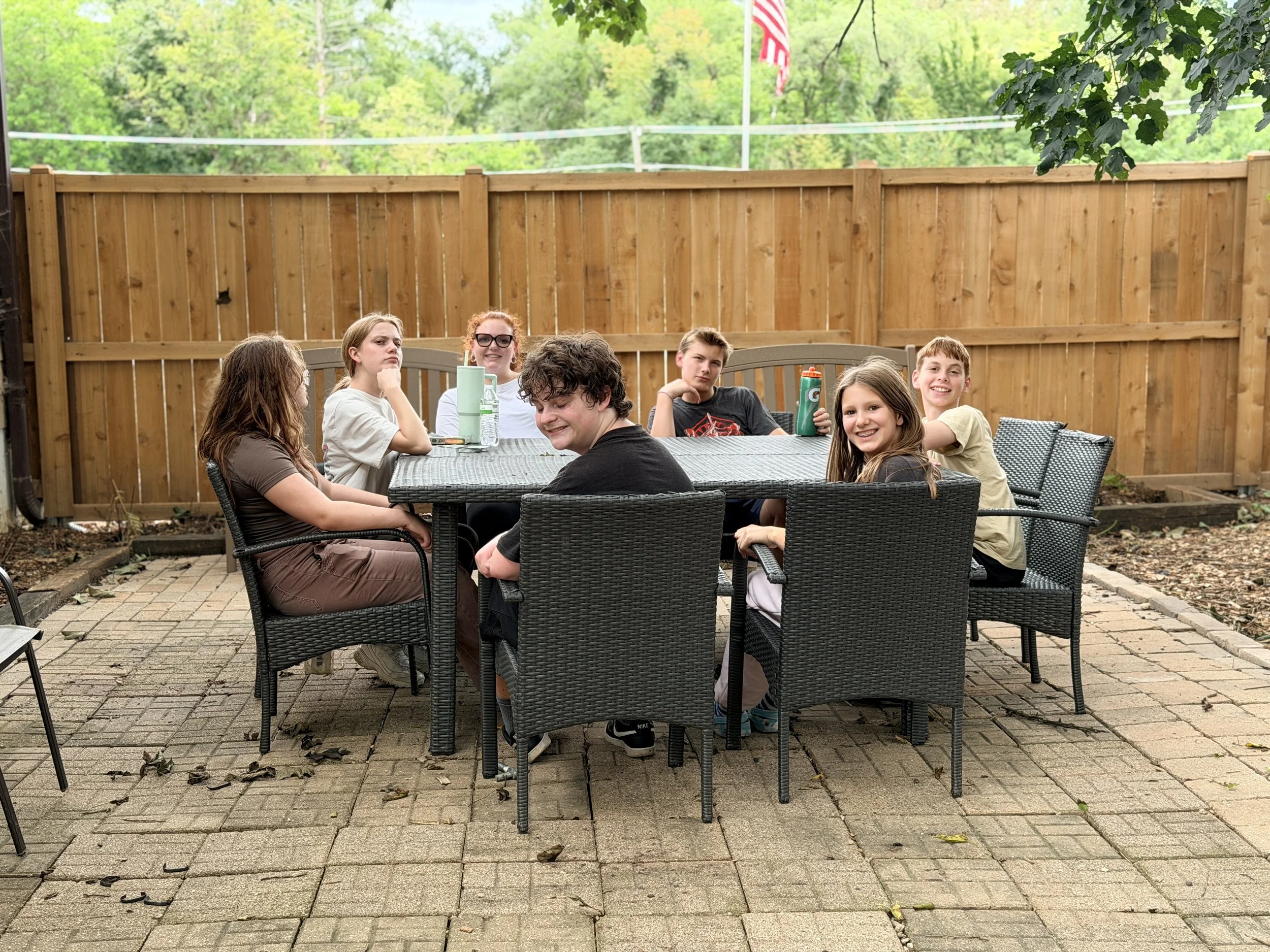 A group of seven young people sitting around a rectangular outdoor table in a backyard with a wooden fence and trees in the background. They appear to be enjoying themselves, with some smiling and others looking at the camera.
