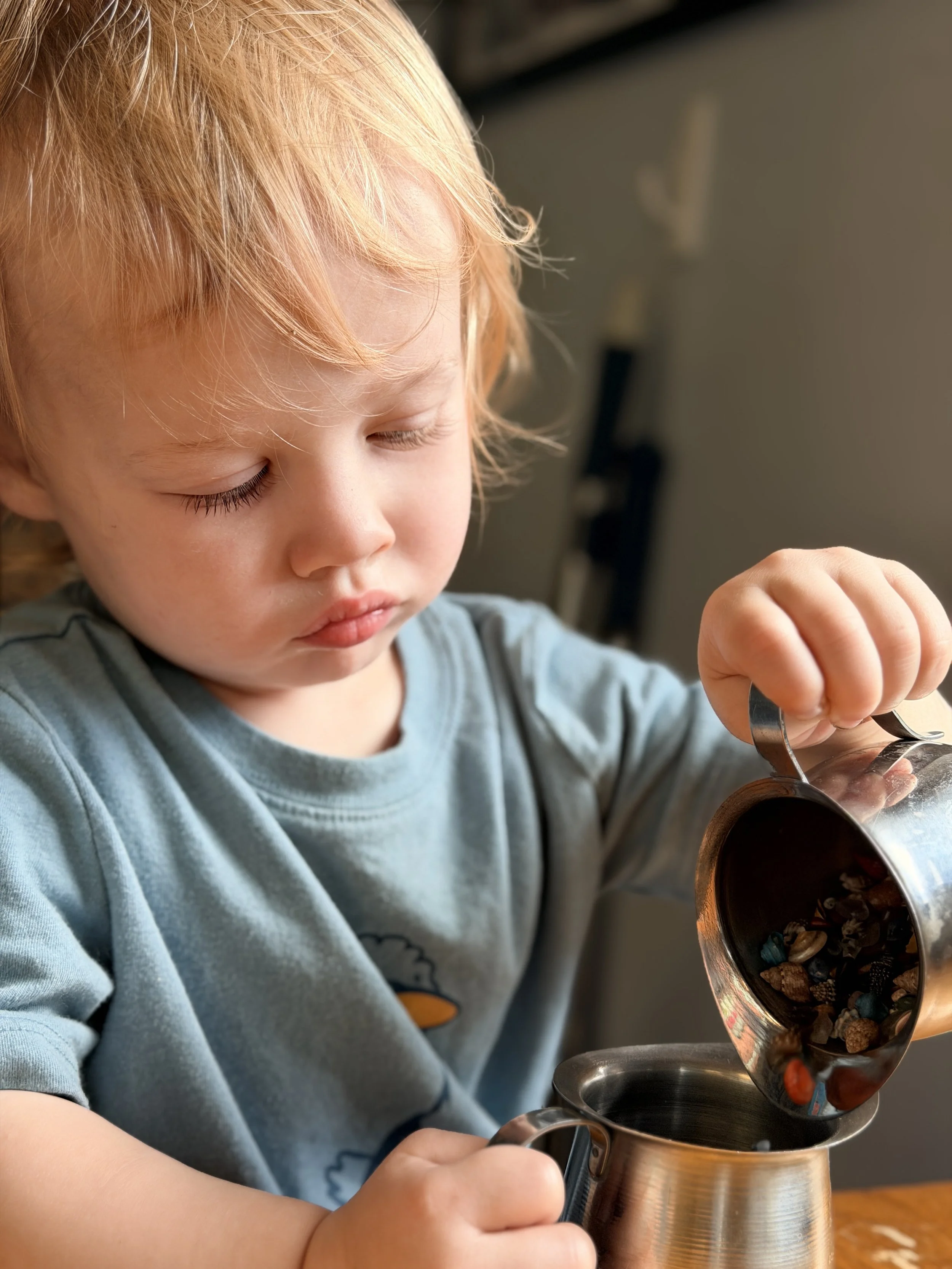 A young child with blond hair and a blue shirt pouring cereal from a metal pitcher into a bowl.