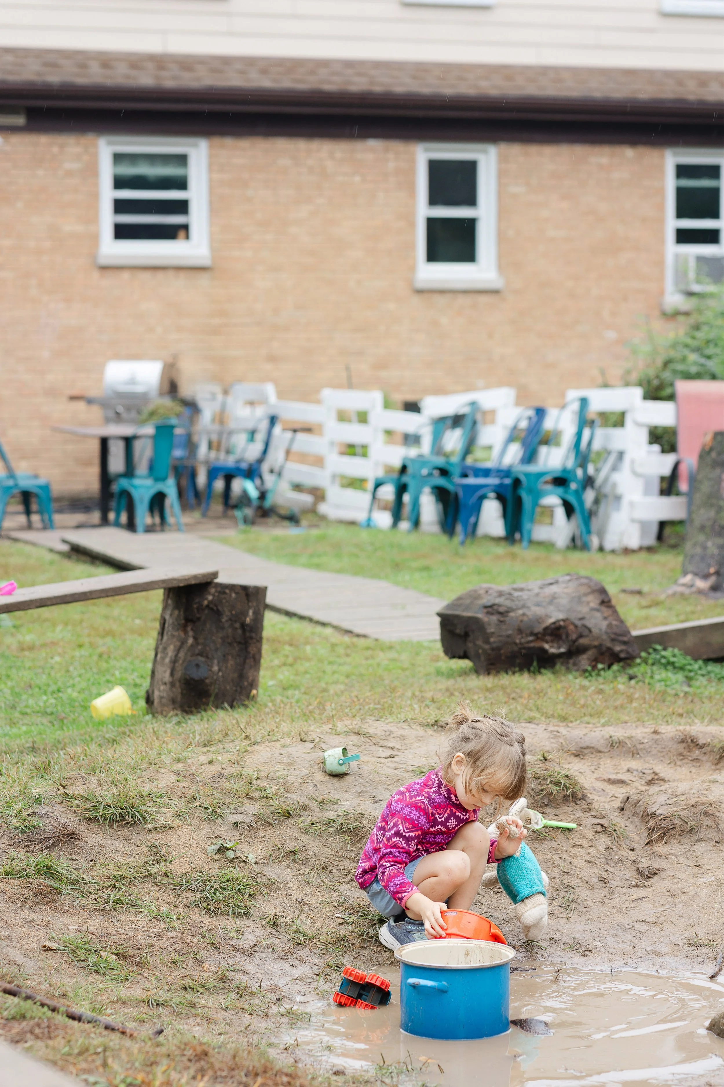 Young girl in pink patterned jacket playing with toys in a muddy puddle outside in a backyard, with outdoor furniture and a brick house in the background.