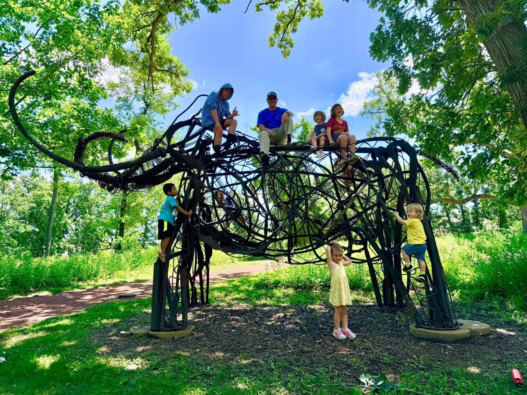 Children and adults playing on a large, tree-shaped metal playground structure in a sunny park with green trees and grass.