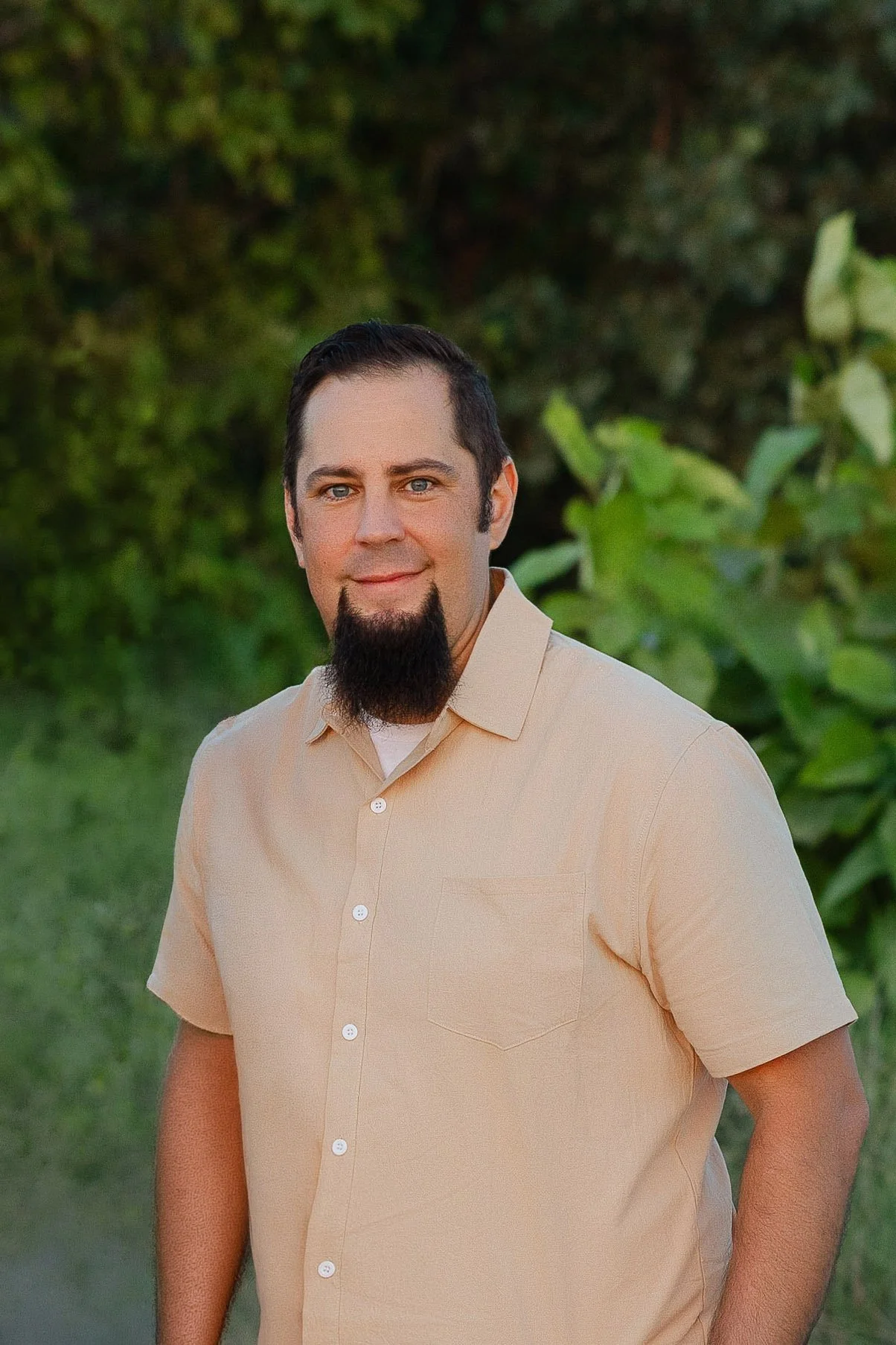 A man with short dark hair, a goatee, and blue eyes wearing a light beige short-sleeved button-up shirt, standing outdoors in front of green foliage.