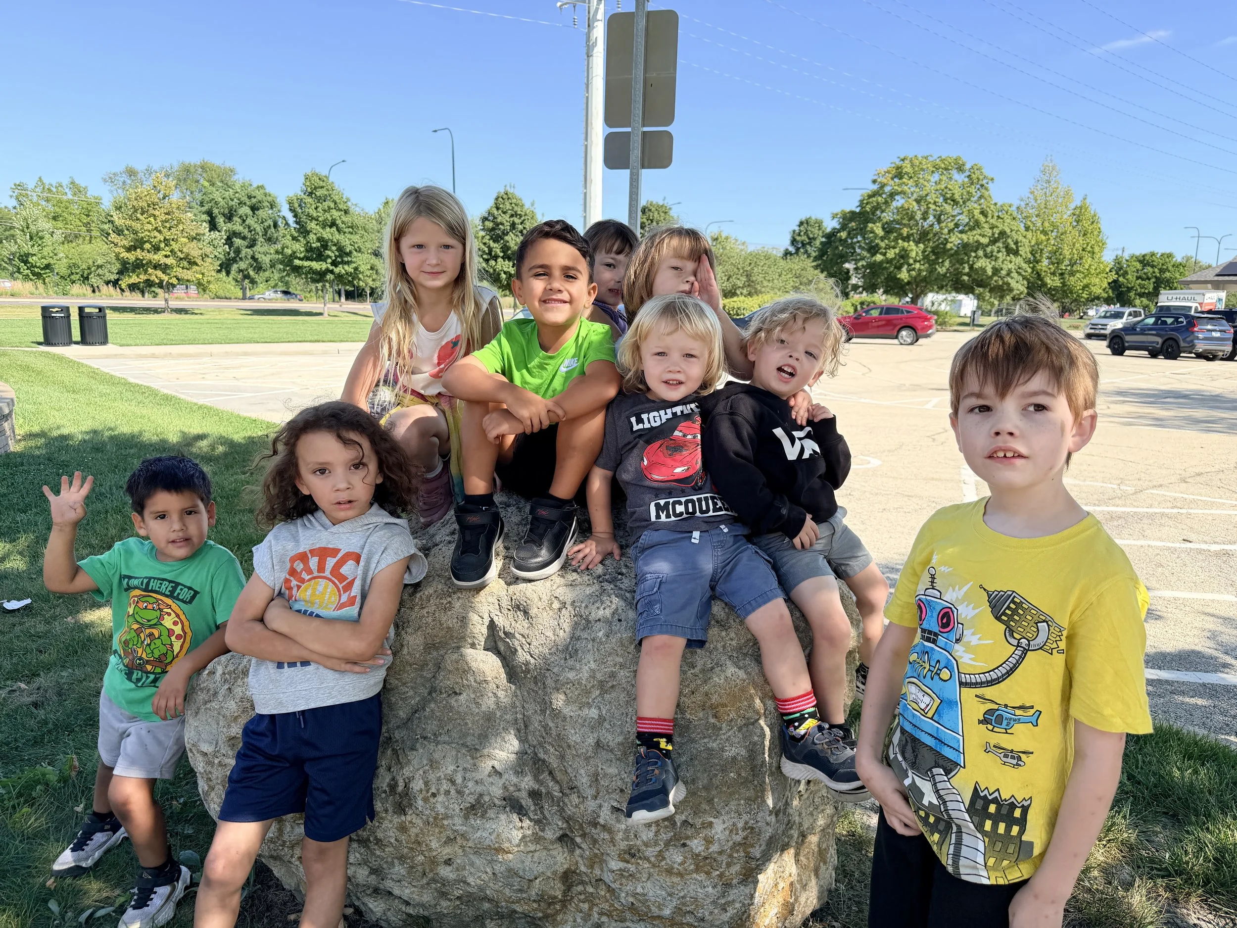 Group of children posed around a large rock in an outdoor setting, some sitting on it and others standing nearby, with trees and parking lot in the background on a sunny day.