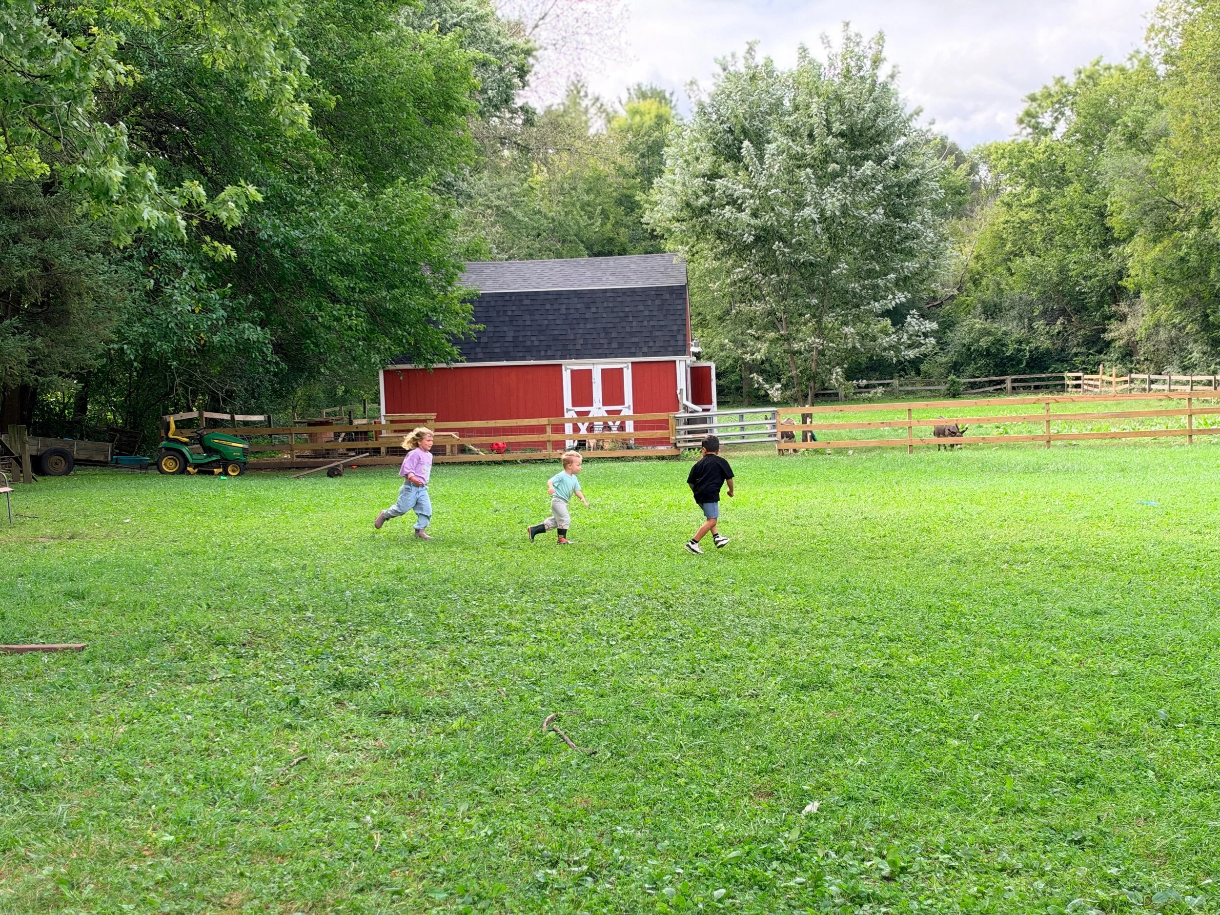 Children playing on a grassy field near a red barn surrounded by trees.