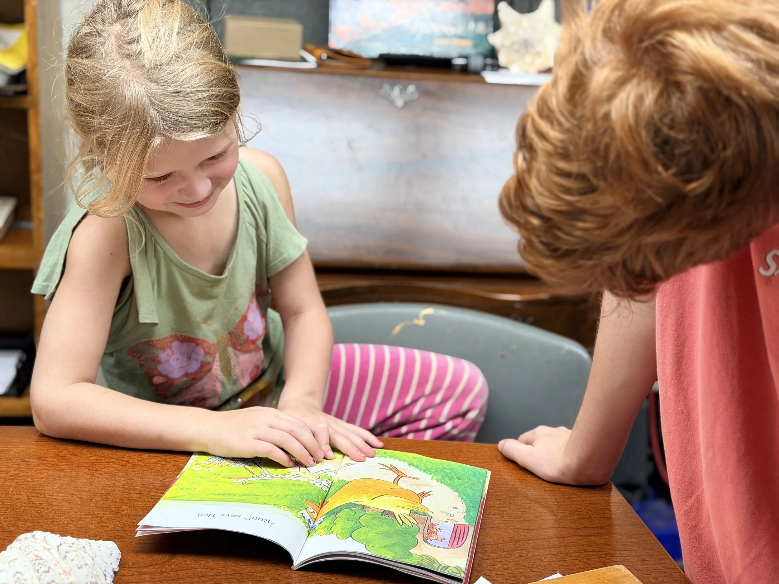 A young girl with blonde hair and a green shirt is reading a colorful children's book, smiling. She is sitting at a wooden table with a person with red hair. The background includes shelves and a map.
