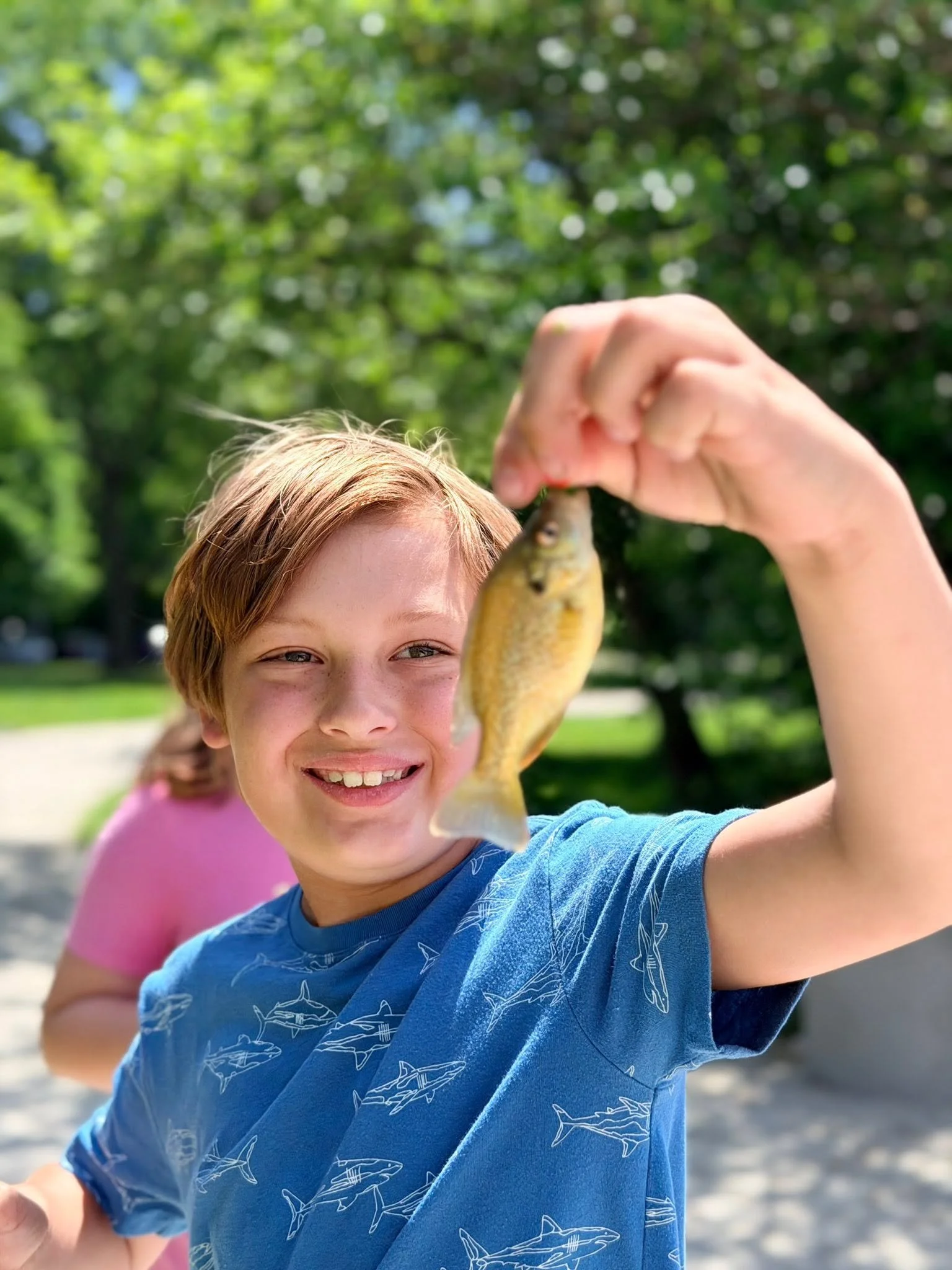 Young boy smiling and holding a small fish with his hand outdoors, with a girl in a pink shirt behind him, in a park or natural setting.