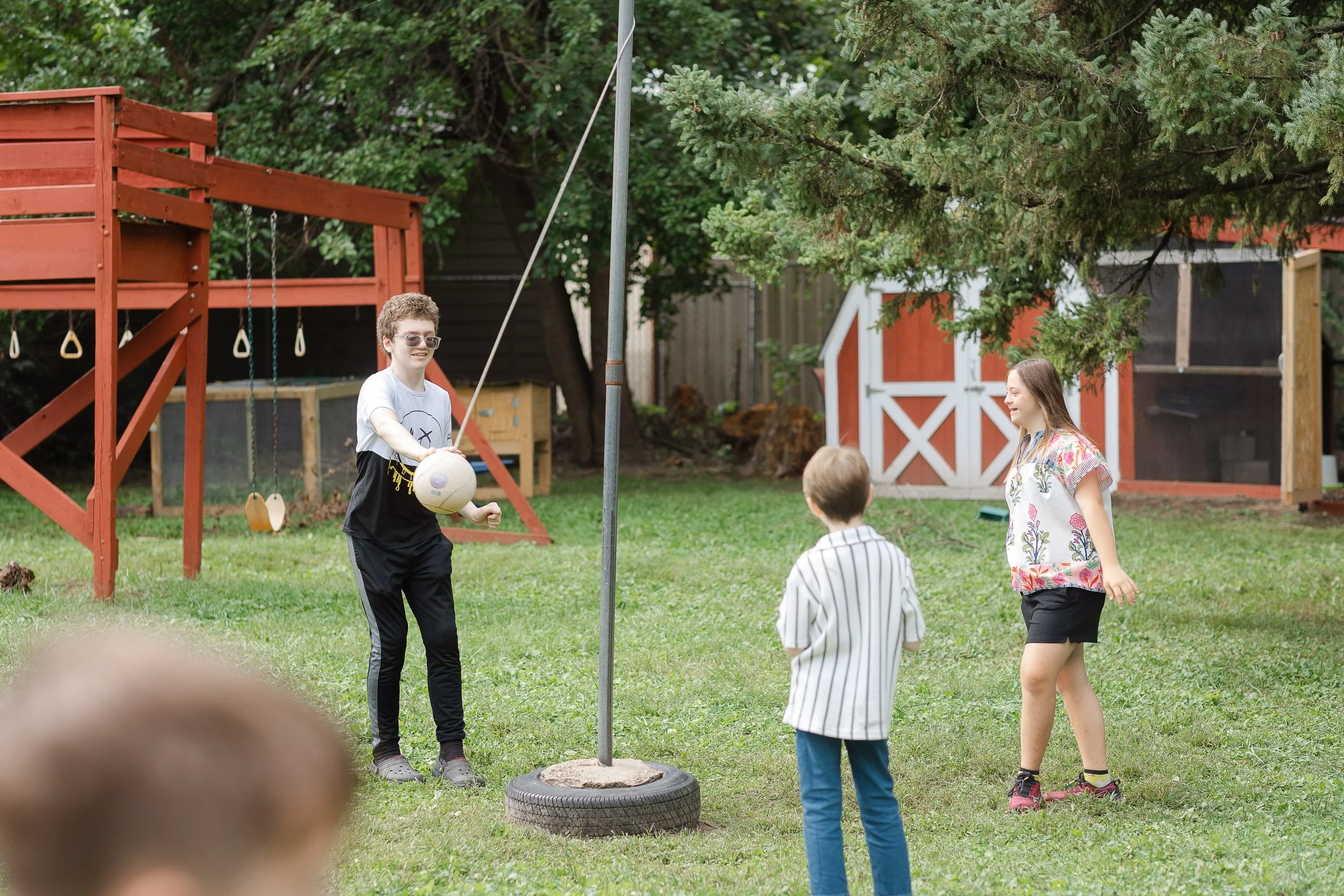 Children playing a game outdoors in a backyard with trees, a red wooden play structure, a shed, and a tire on the ground.