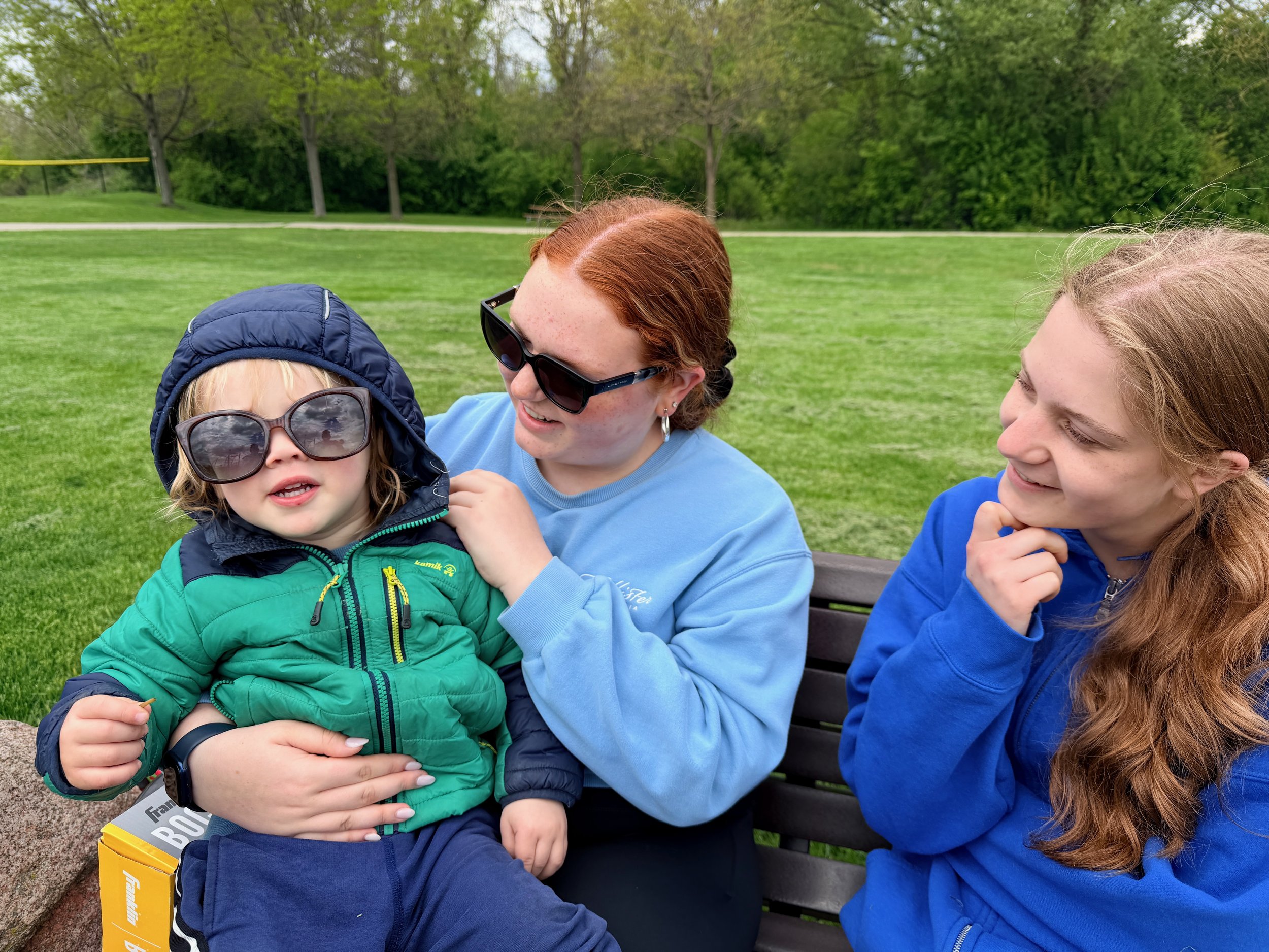 A young girl with curly blond hair and wearing sunglasses is sitting on a park bench holding a slightly older woman with red hair and glasses, who is smiling and looking at her. Another woman with long curly brown hair and a blue hoodie is to the sid