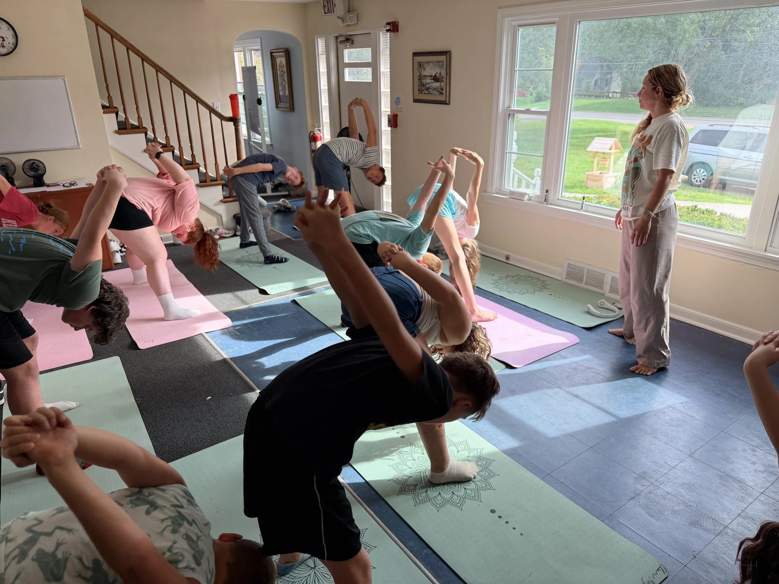 A group of children participating in a yoga class indoors, with some children stretching forward on their mats and others bending backwards, while an instructor oversees the activity near a large window with a green outdoor view.