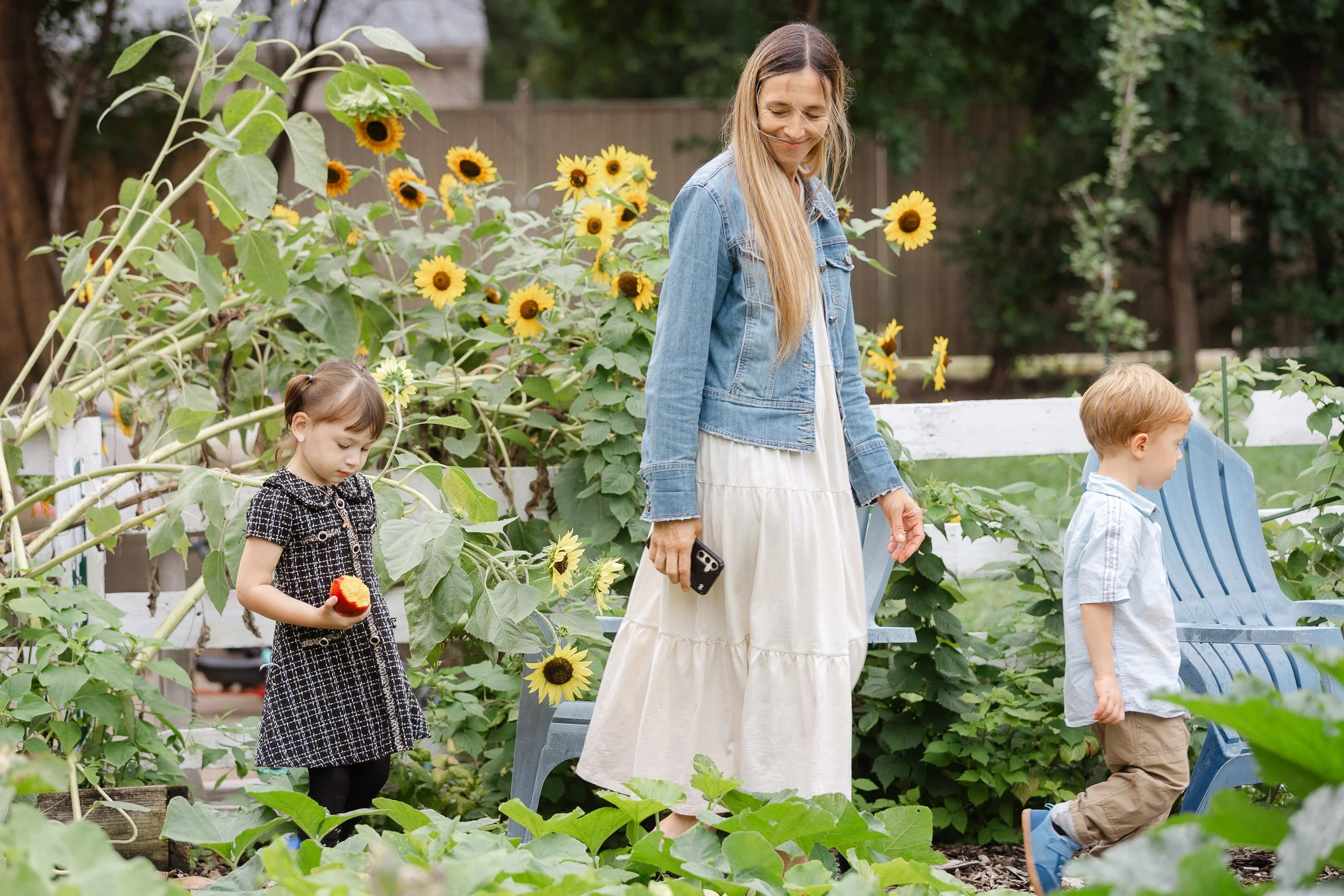 A woman and two children in a garden with sunflowers, the woman is holding a phone and smiling while the children are engaged in gardening activities.