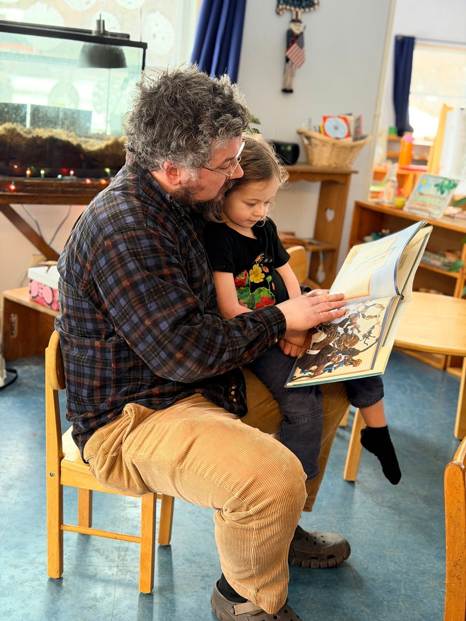 An older man with curly gray hair and glasses reading a book with a young girl with curly brown hair, both sitting on chairs in a room filled with colorful toys and books.