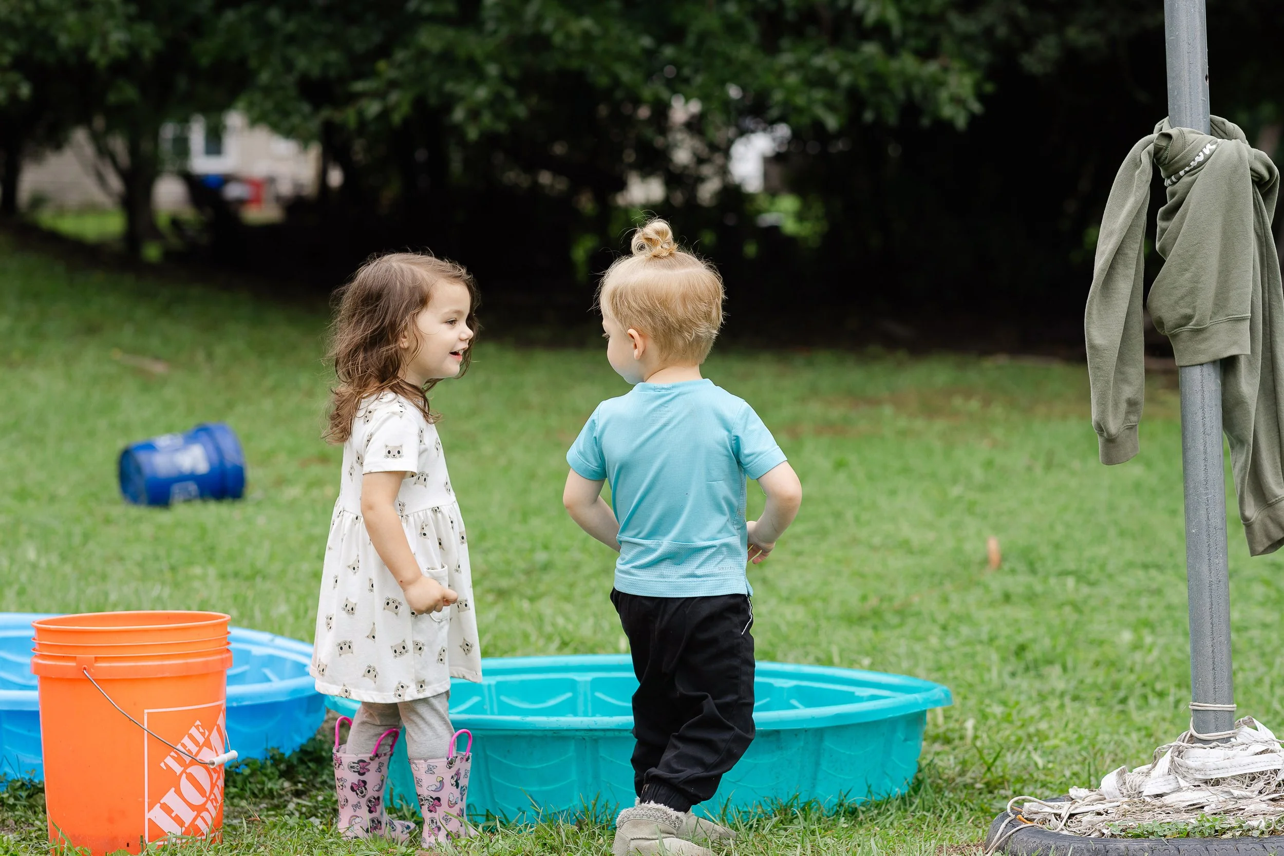Three children, two girls and one boy, are standing near an inflatable pool outdoors, talking and smiling. There are orange and blue buckets nearby, and a skirt and shoes are hanging on a clothesline attached to a pole.