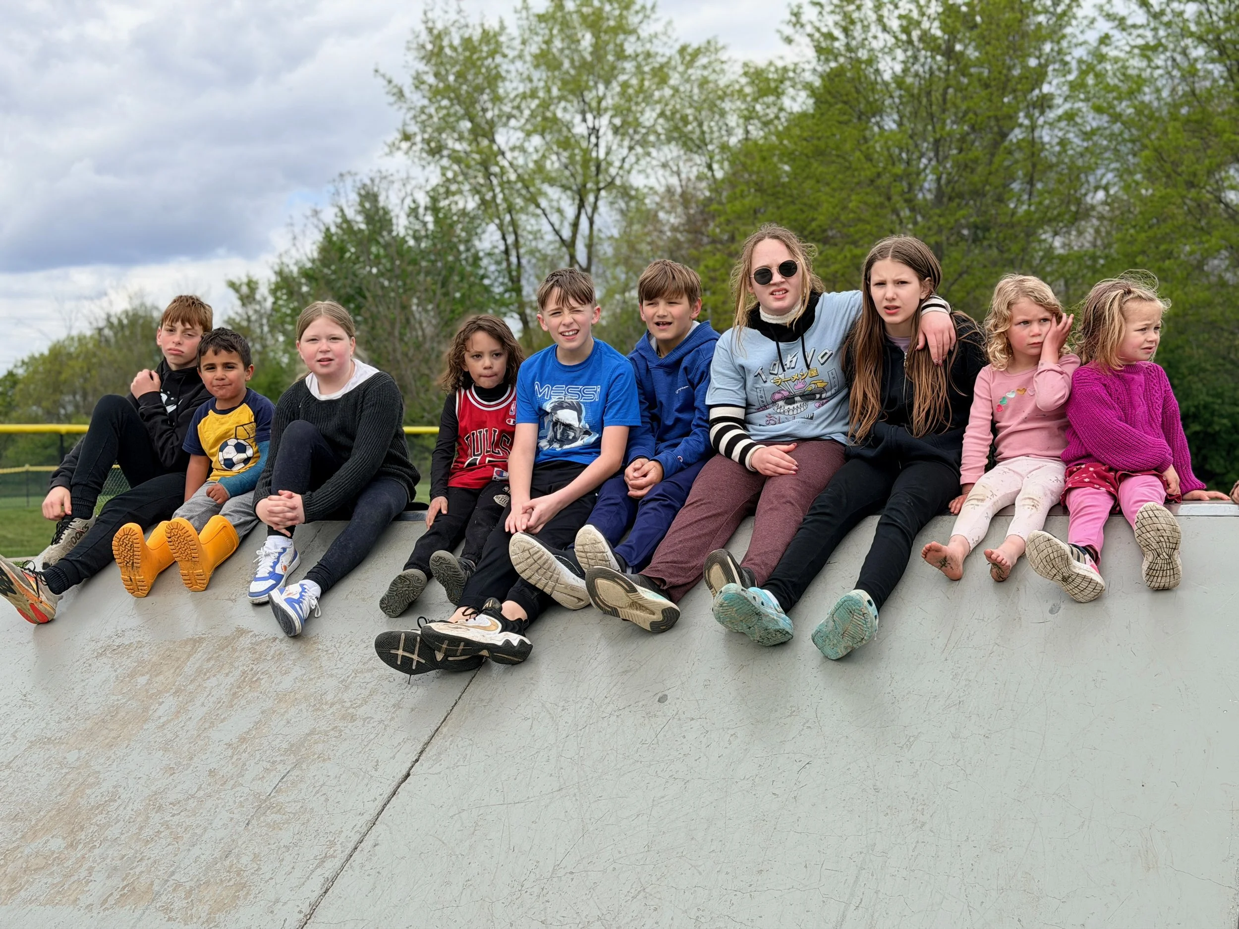A group of children sitting on the edge of a skate ramp outdoors, with trees and a cloudy sky in the background.