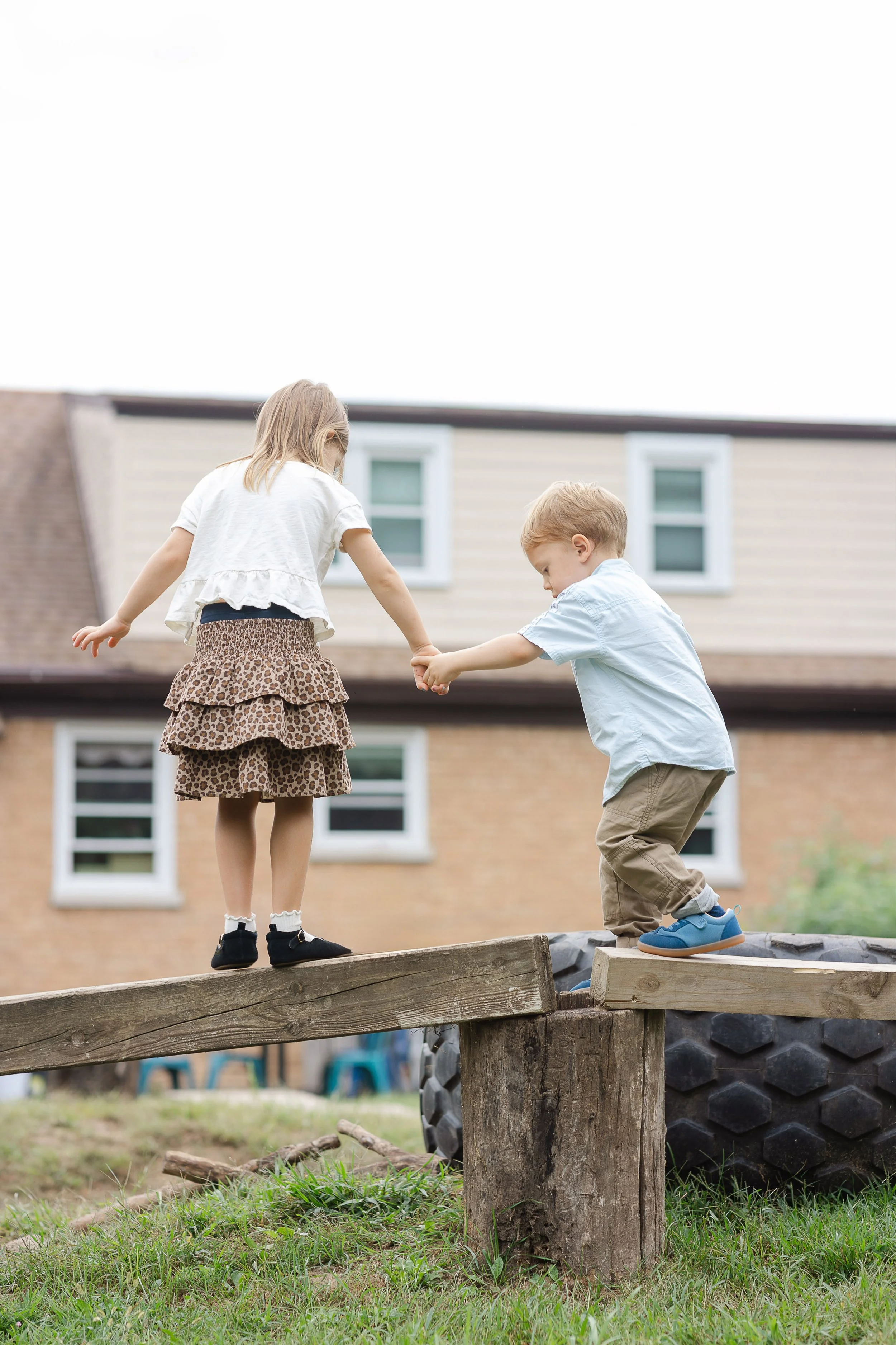 Two children, a girl with blonde hair and a boy with light brown hair, are holding hands and balancing on a wooden beam in a backyard with a house in the background. The girl wears a white top, leopard-print skirt, and black shoes, while the boy wear