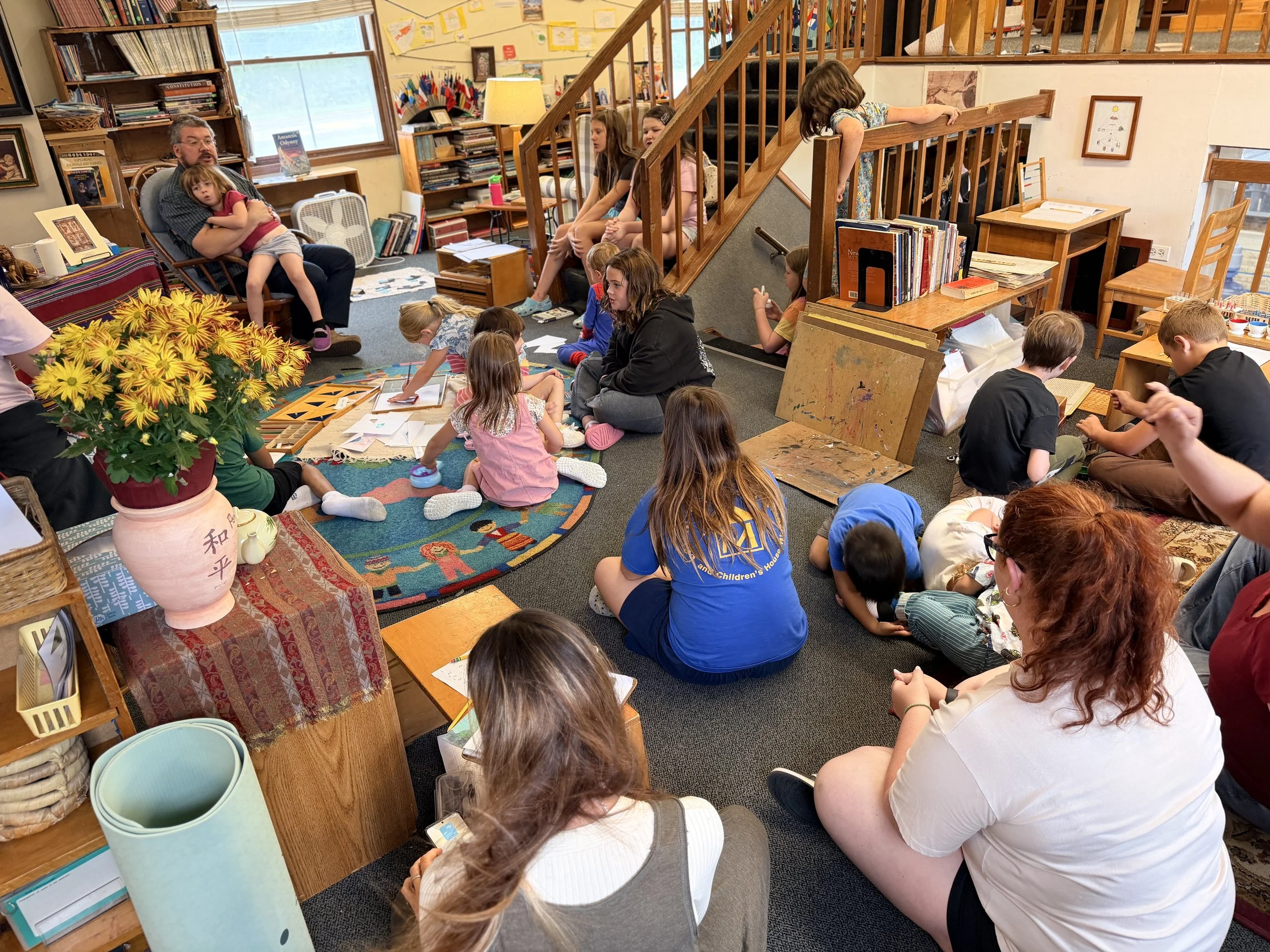 A group of children and adults gathered inside a cozy bookstore or library, sitting on the floor and chairs, listening to a man holding a young girl, while bookshelves and artwork decorate the background.