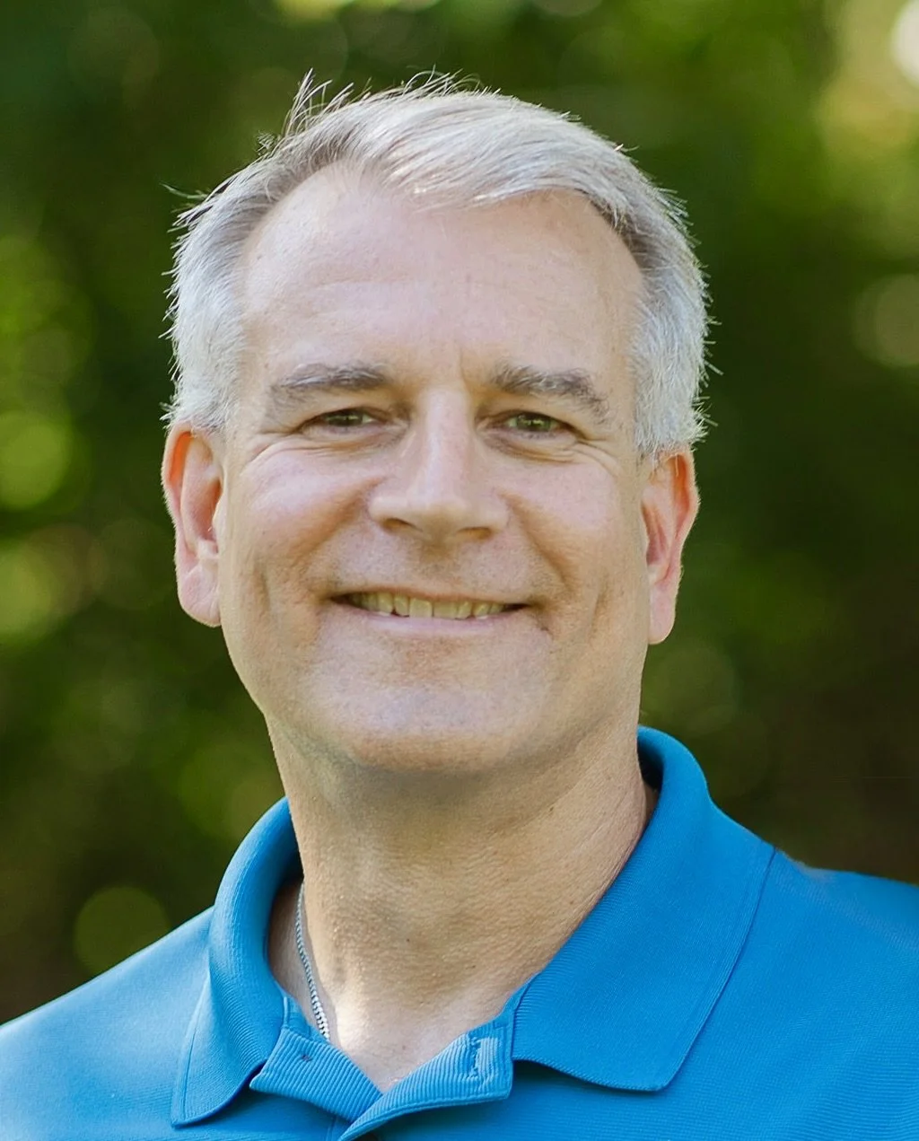 A middle-aged man with short gray hair and light skin, smiling outdoors with a blurred green background, wearing a blue collared shirt.