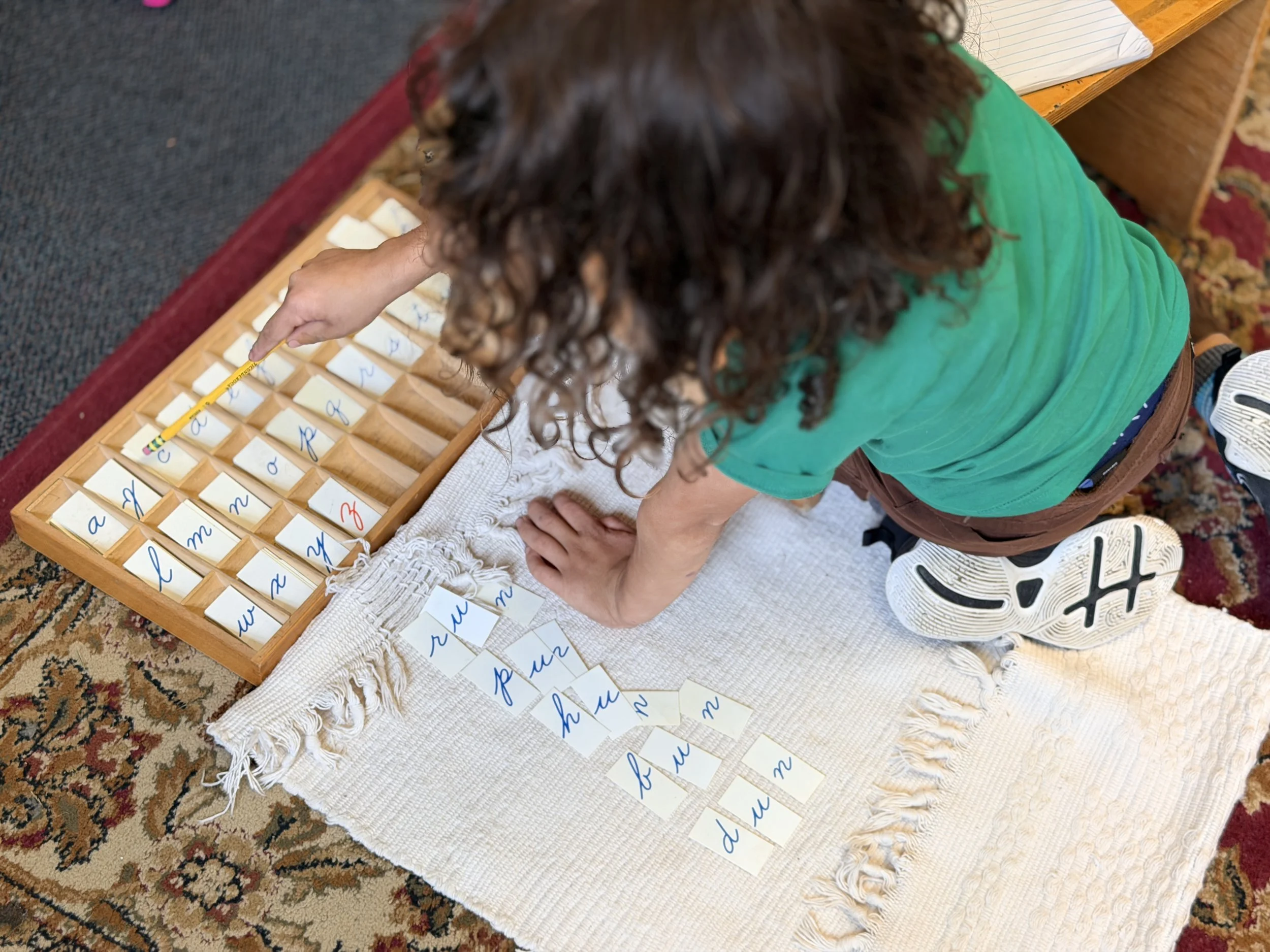 A young child with curly hair, wearing a green shirt, is pointing to a tray with slices of paper with cursive letters on them, on a table covered with a white cloth. The child is kneeling on a patterned rug.