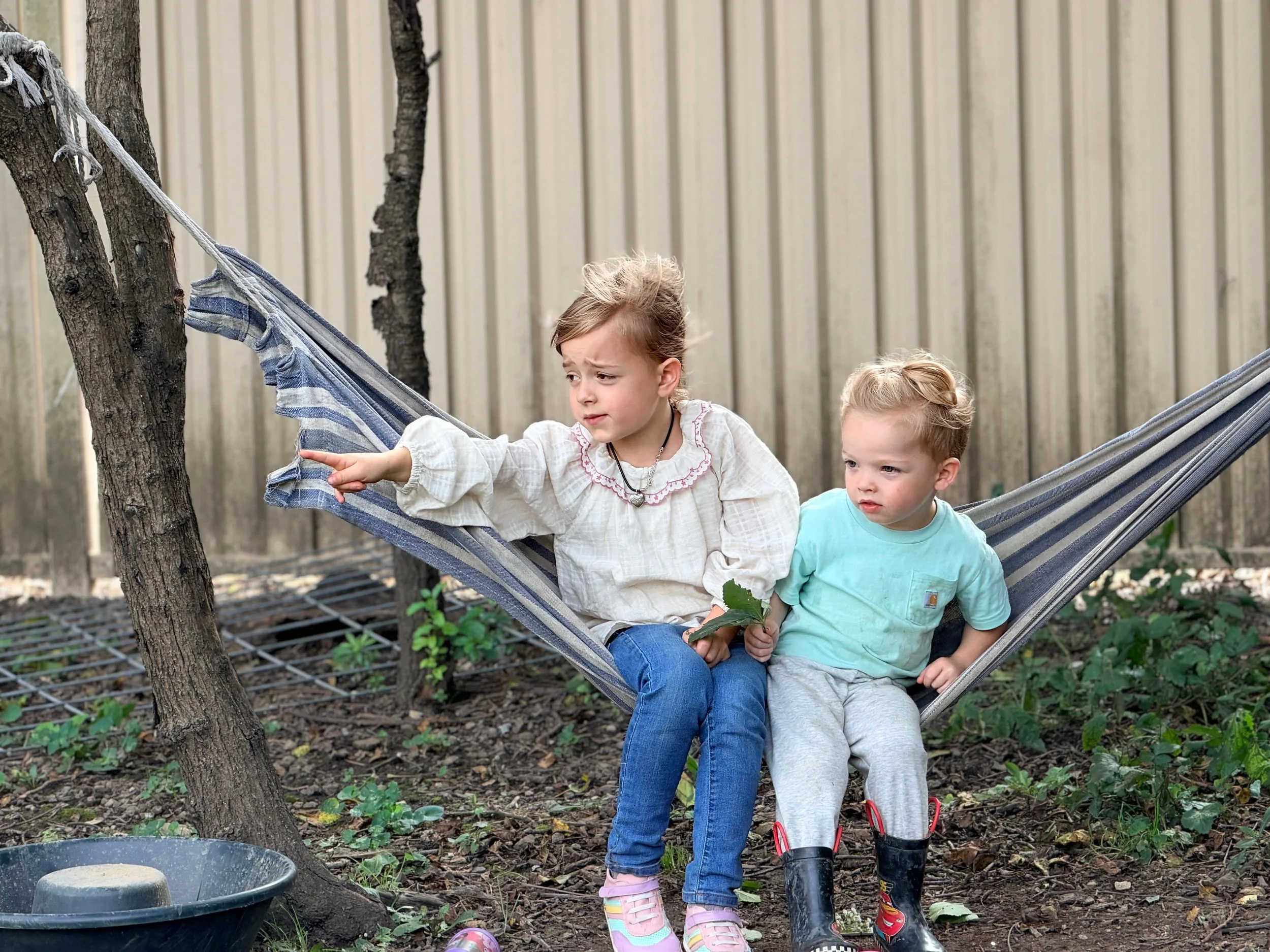 Two young girls sitting on a hammock in a backyard, holding a leaf and pointing at something. The girl on the left is wearing a white dress with embroidery, and the girl on the right is wearing a light blue shirt. There is a sandbox with a stone on t
