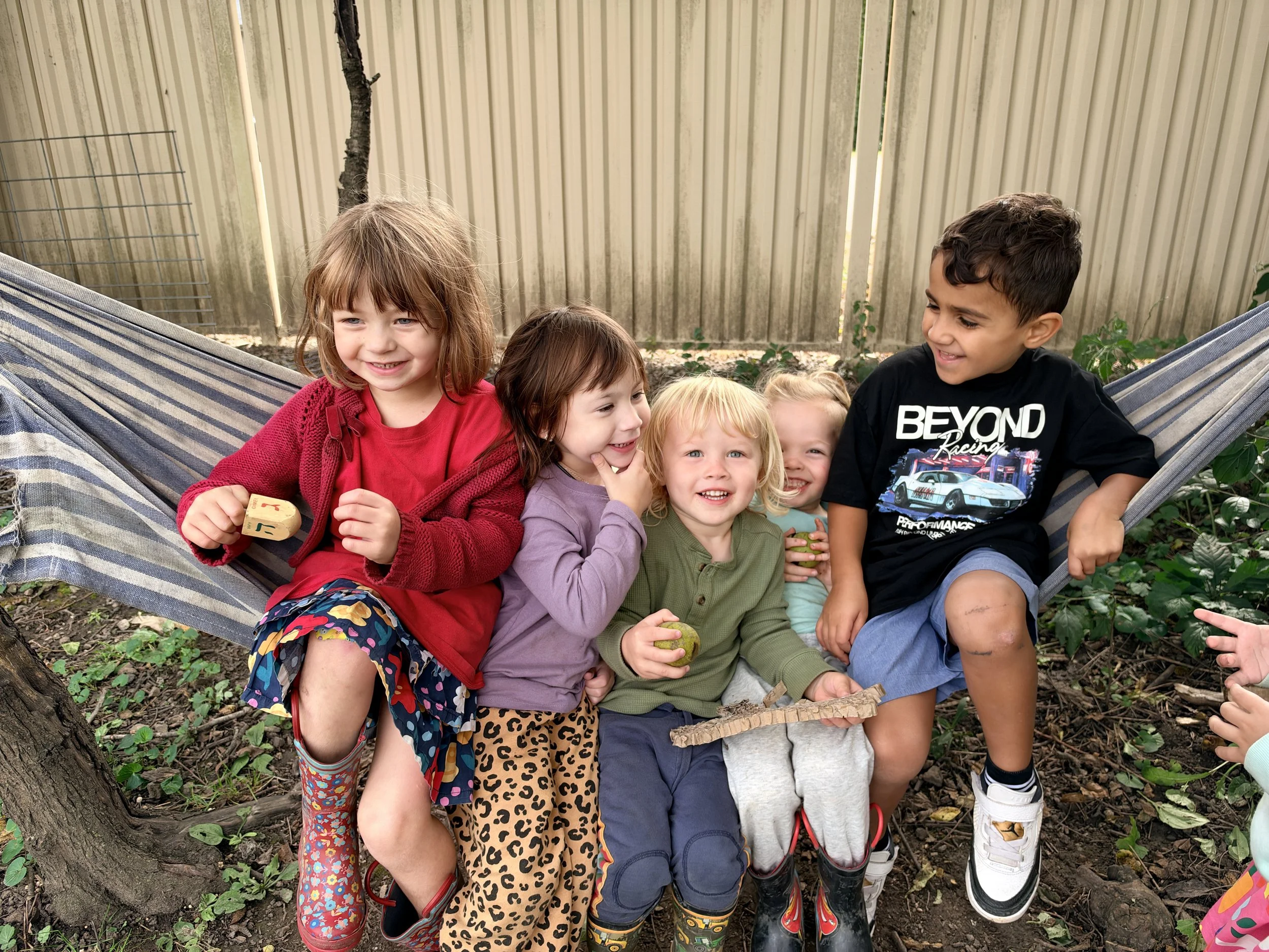 Six children sitting together outdoors on the ground near a tree, smiling and looking at each other, in a backyard with a wooden fence in the background.