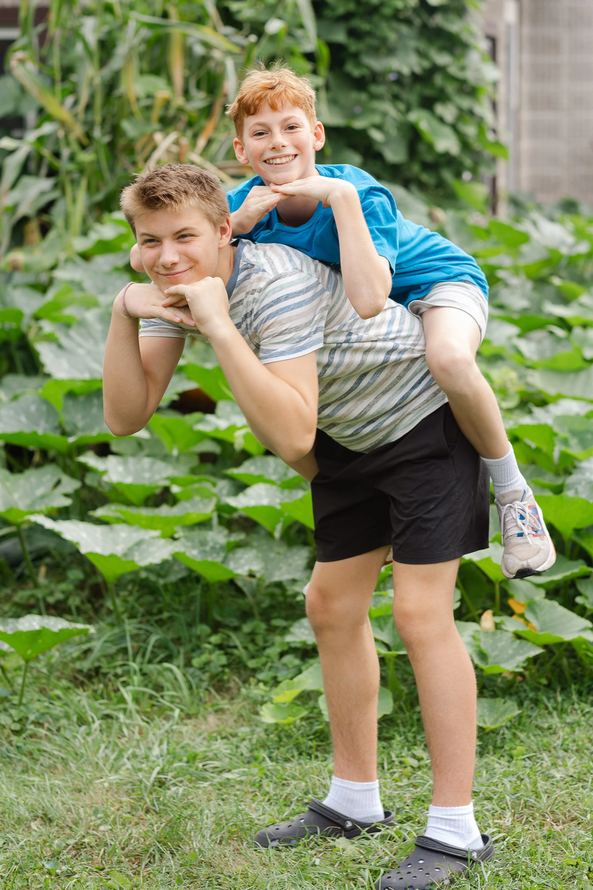 Two boys playing outside in a garden, with one carrying the other on his back. They are smiling and surrounded by green plants and grass.