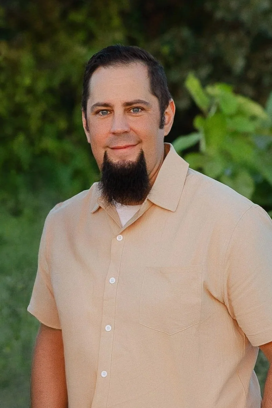 A man with short dark hair, a goatee, and blue eyes wearing a light beige short-sleeved button-up shirt, standing outdoors in front of green foliage.