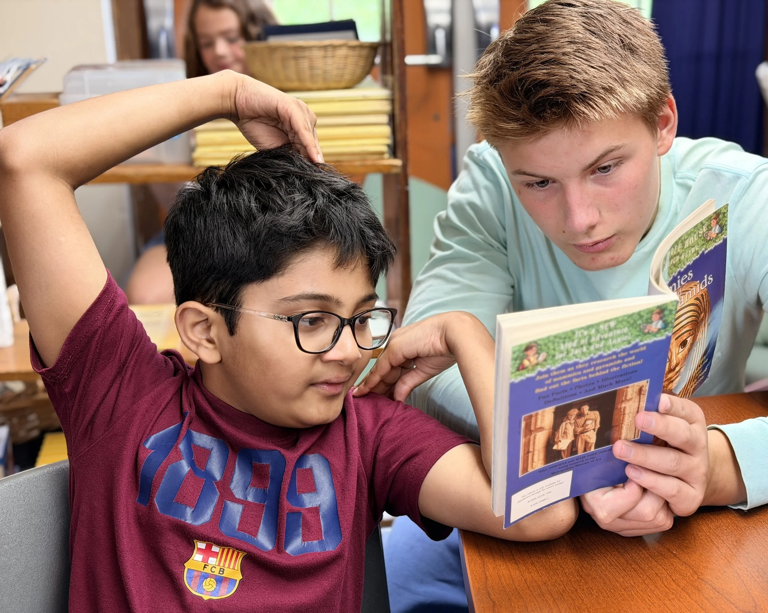Two boys are reading a book together while sitting at a table, with a girl behind them looking at a computer.