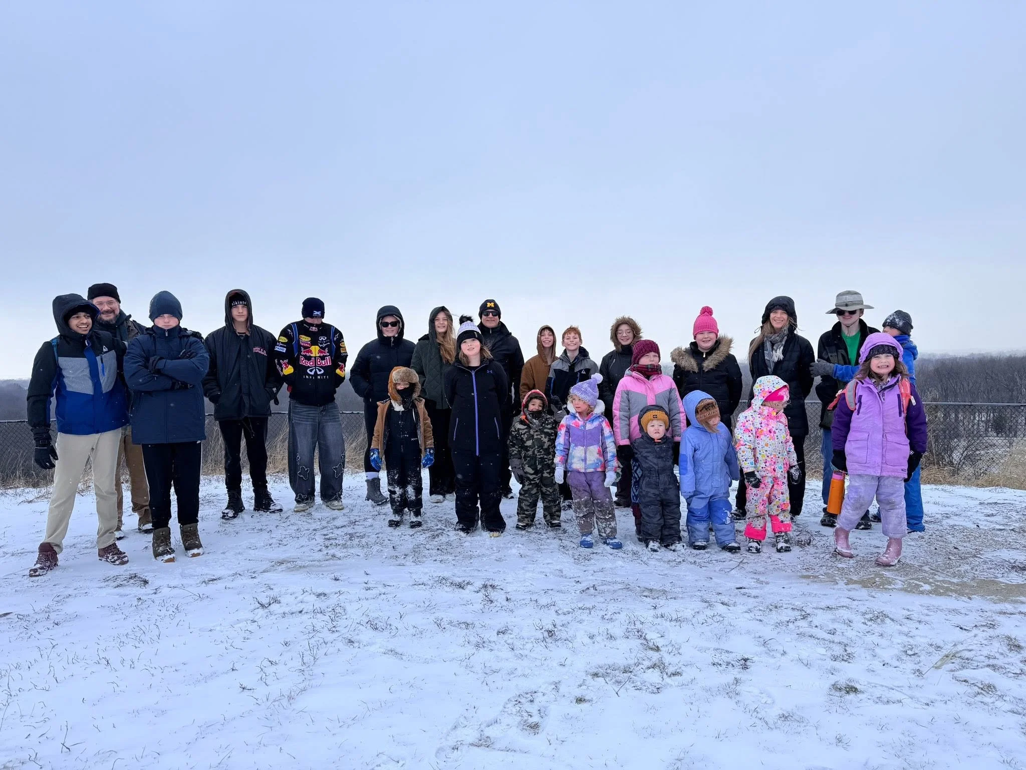 A group of children and adults standing outdoors in a snowy field on a cloudy day, dressed in winter clothing.