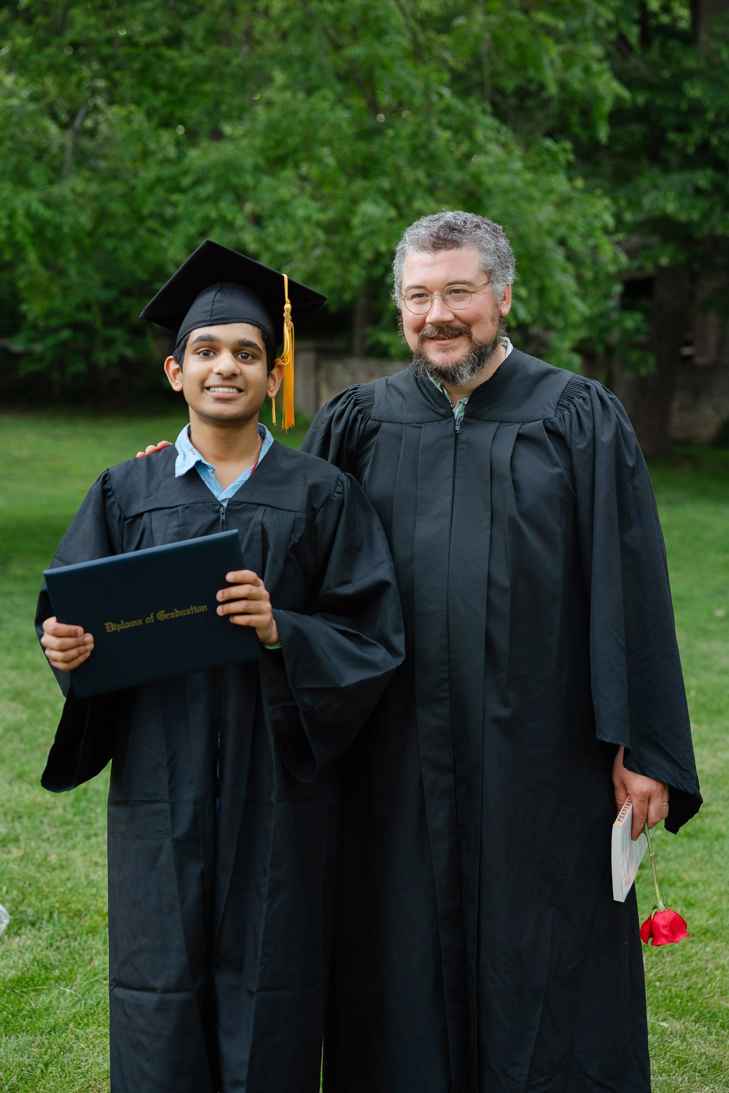 A young man in a black graduation gown and cap holding a diploma, standing next to an older man in academic robes, outdoors with green trees in the background. The young man is smiling, and the older man has a beard, glasses, and is smiling as well.