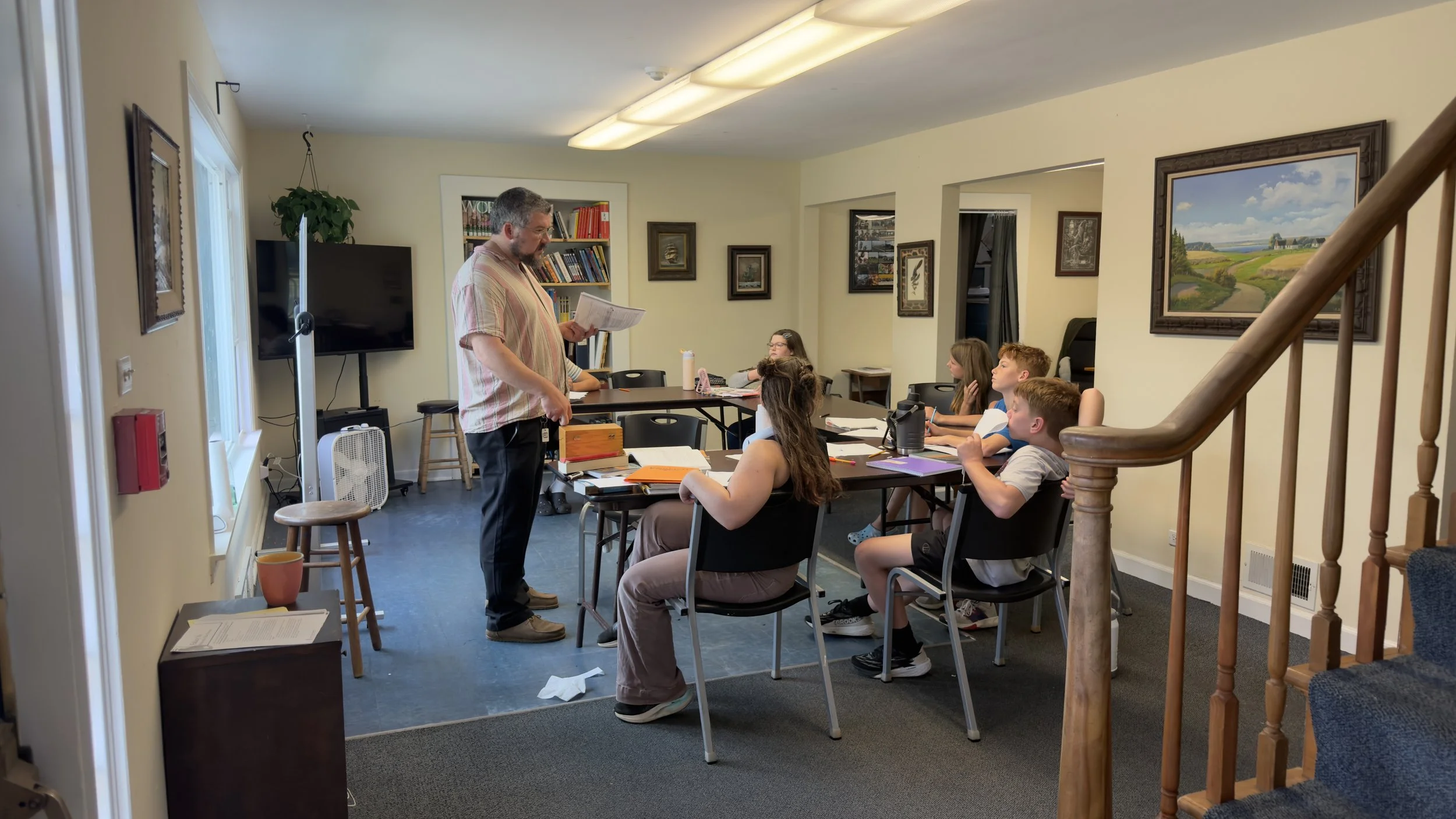 A classroom with a teacher standing in front of six students who are seated at a long table. The teacher appears to be explaining something from a book or paper. The room has artwork on the walls, a large television, and a window letting in natural l