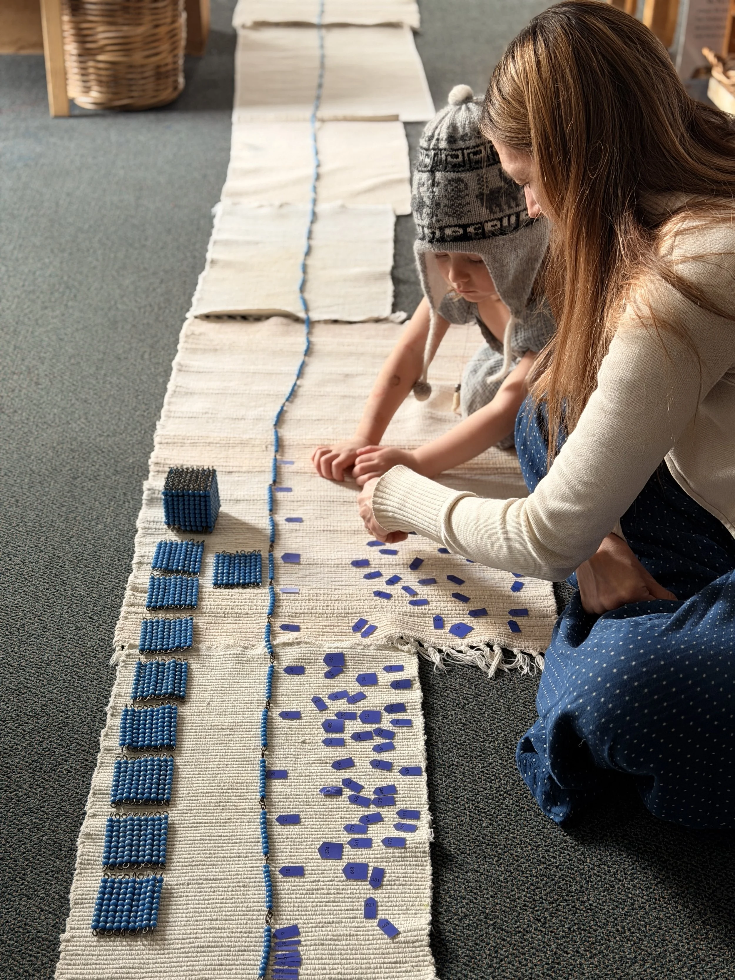 A woman and a young boy sit on the floor, using blue and black alphabet tiles to spell out words on a long white woven rug.