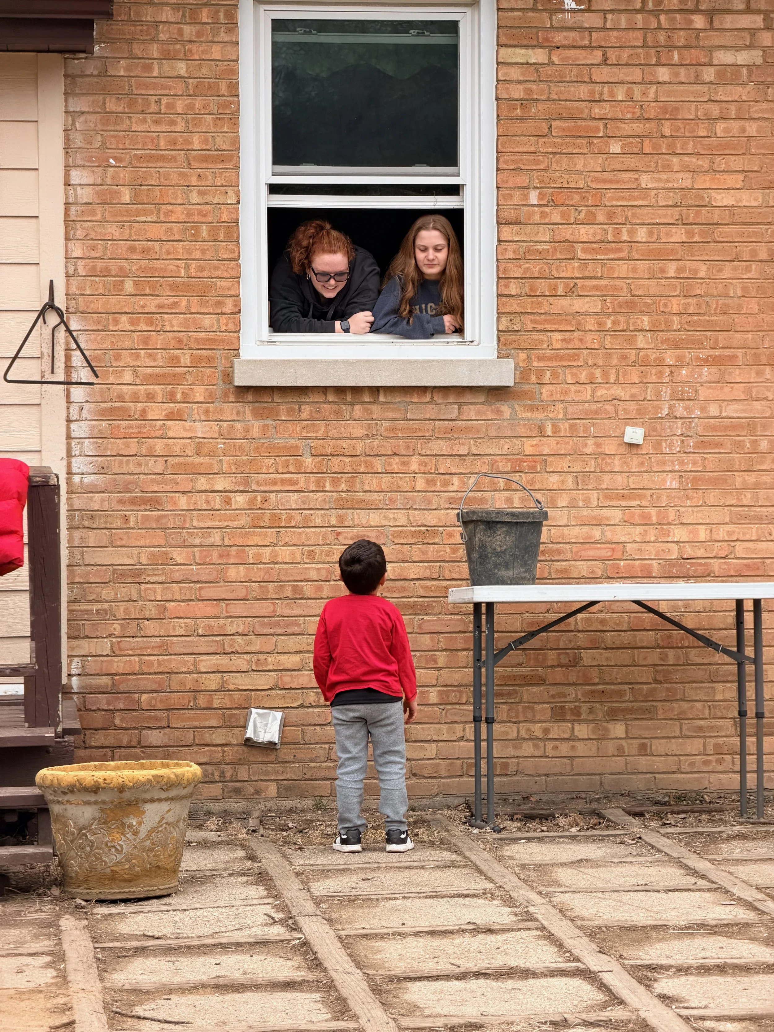 A young boy with short dark hair wearing a red sweatshirt and gray pants standing outside on a patio, looking at two women by a window of a brick house. The women are leaning out of the window, one with curly hair and glasses, the other with long str