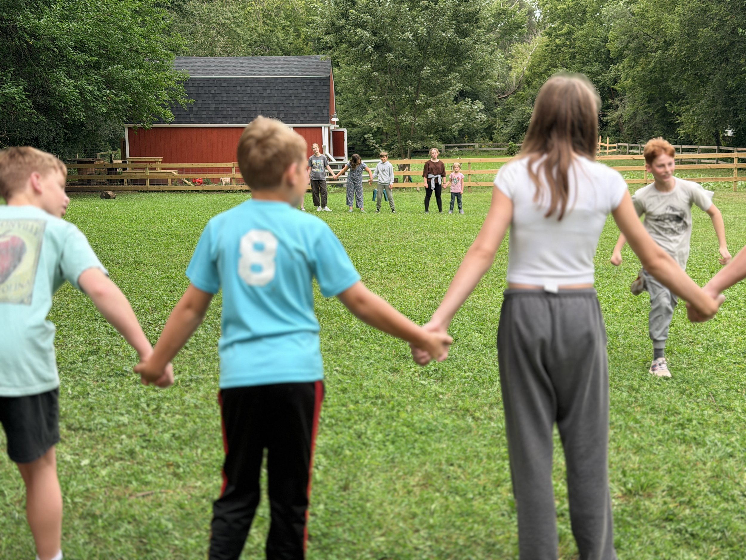 Children holding hands, forming a circle outdoors on a grassy area with trees and a red barn in the background.