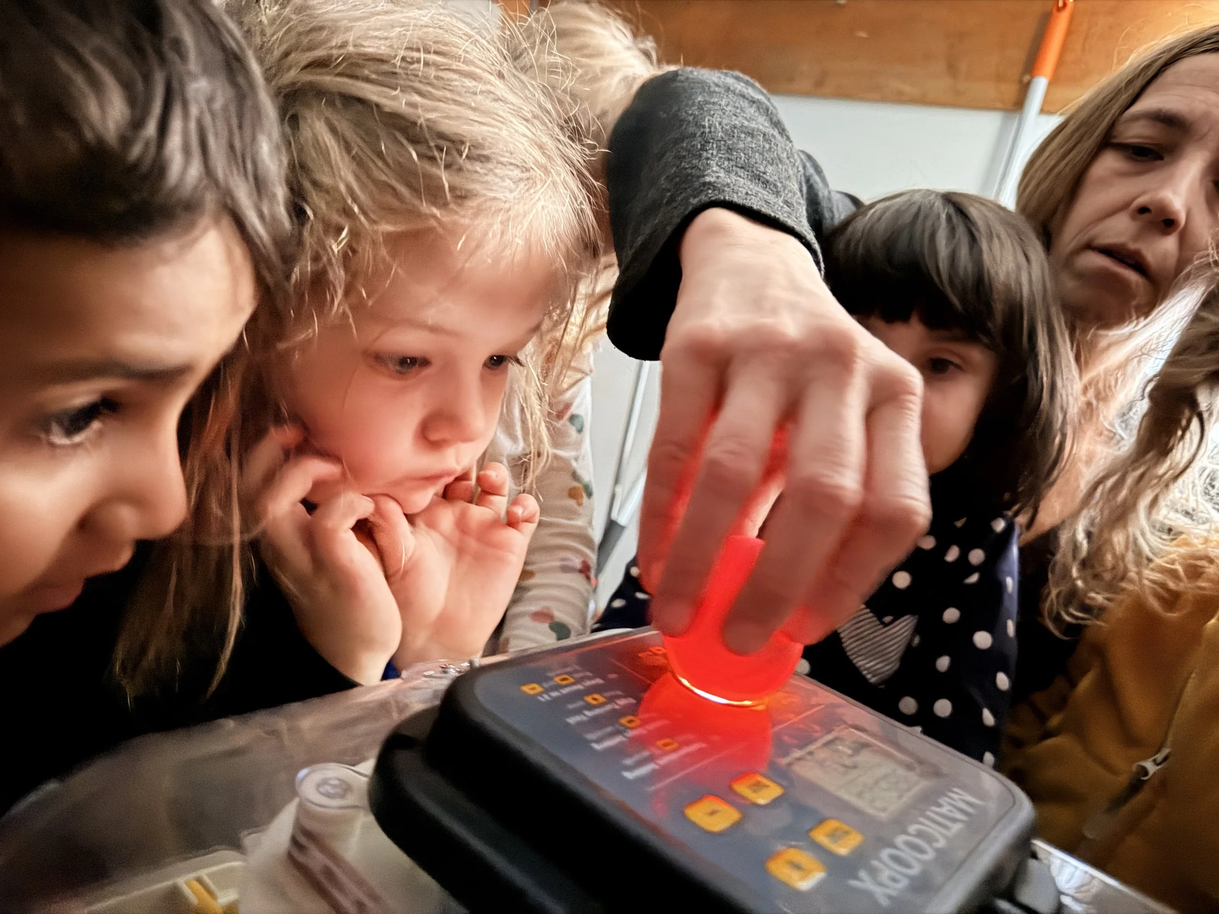 Close-up of three children and an adult looking at a detective board game on a table, with a red plastic magnifying glass and a tablet nearby.